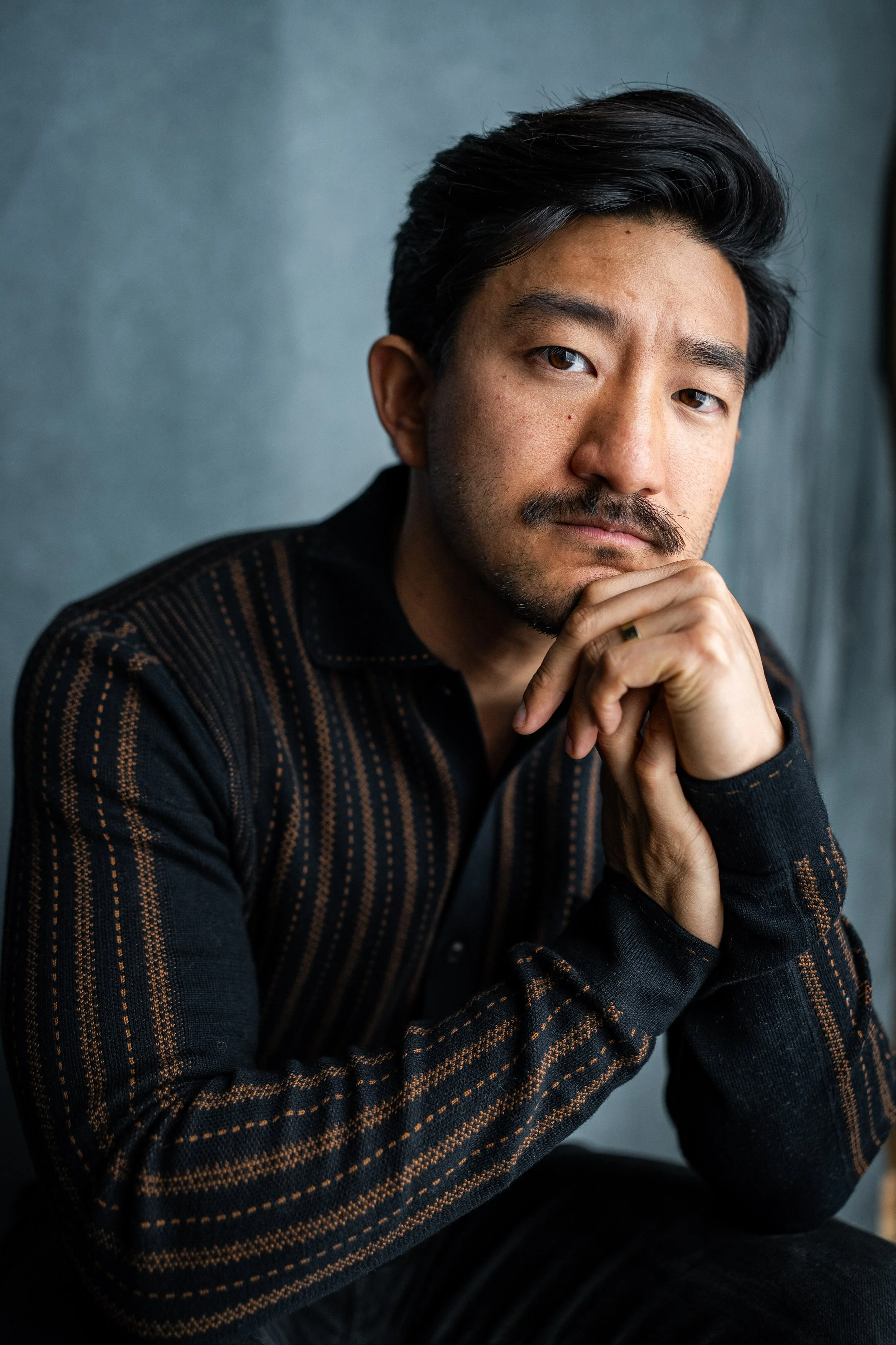 Portrait of a young Asian man with dark, wavy hair, wearing a dark blazer over a white shirt, against a plain background.
