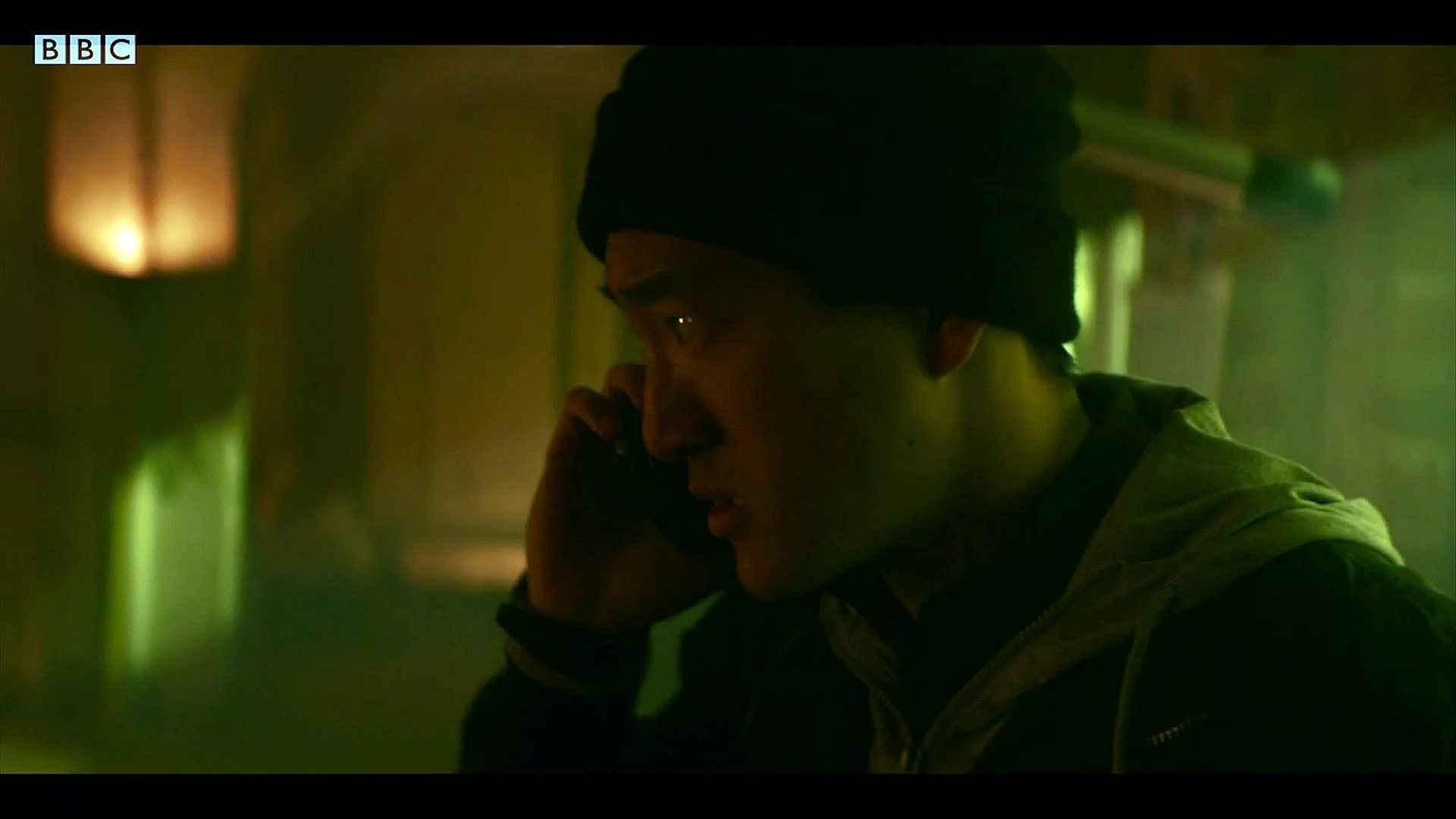 A young person speaking on a mobile phone in a dimly lit, greenish-hued room, wearing a black beanie and a light-colored zipped jacket.