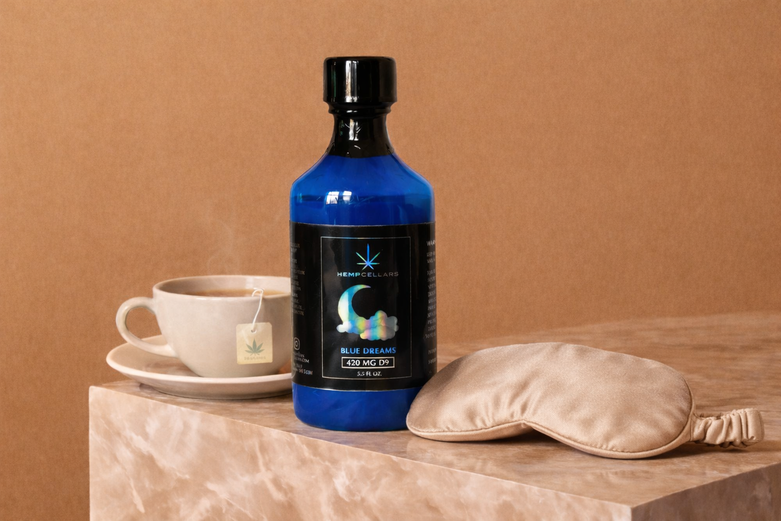 A blue bottle of hemp dietary supplement labeled "Blue Dreams" next to a beige sleep mask and a white cup of tea on a marble surface with a pink background.