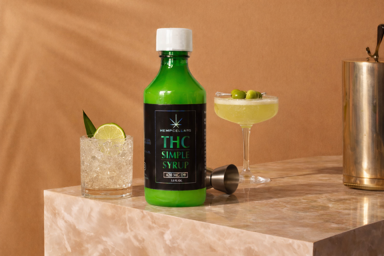 A bottle labeled 'THC Simple Syrup' with a cannabis leaf logo on a marble countertop, accompanied by two cocktails, one with ice and a lemon slice, the other in a martini glass with green olives, and a metal cocktail shaker.