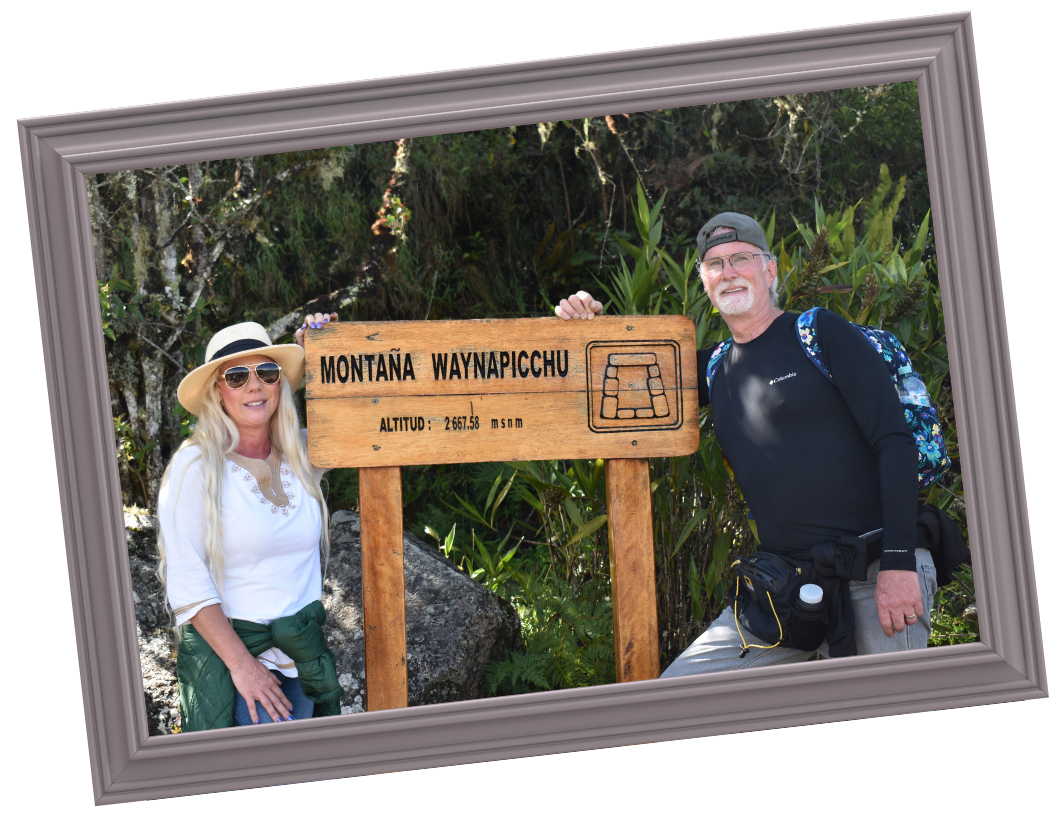 Two hikers, a woman with long blonde hair wearing a white hat, sunglasses, and a white shirt, and a man with glasses, a black cap, and a black shirt, standing outdoors next to a wooden sign that reads 'Montaña Waynapicchu' with an altitude of 2,667.58 meters, surrounded by greenery.