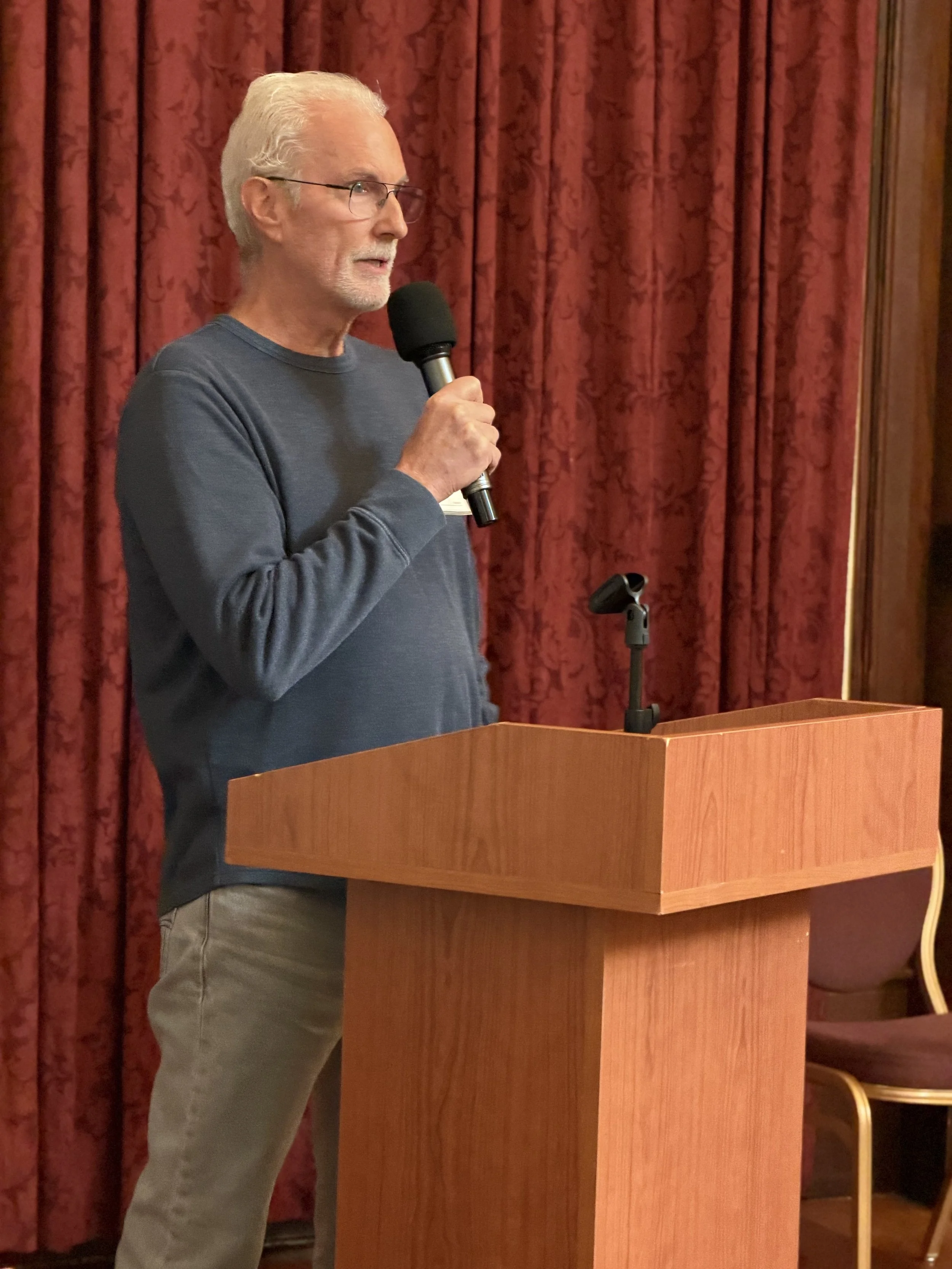 An older man with white hair, glasses, and a beard is standing at a wooden podium holding a microphone, speaking in front of a red curtain.