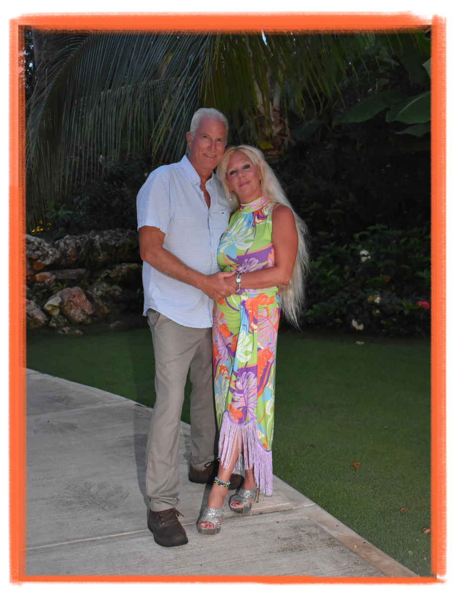 A man and woman standing close together outdoors at sunset, smiling. The man is wearing a light blue short-sleeve button-up shirt and beige pants, and the woman is wearing a colorful, tropical-patterned dress with fringe at the bottom and silver high heels. They are holding hands, surrounded by lush greenery and tropical plants.