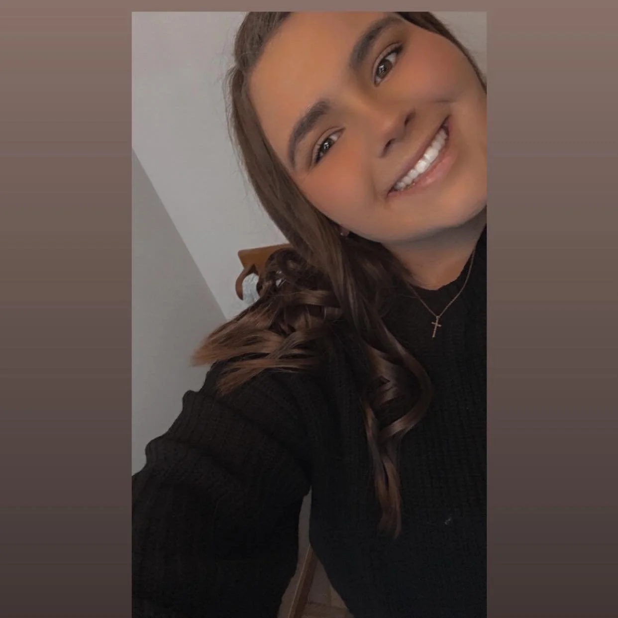 Smiling woman with long wavy brown hair, wearing a black sweater and a cross necklace, taking a selfie indoors.