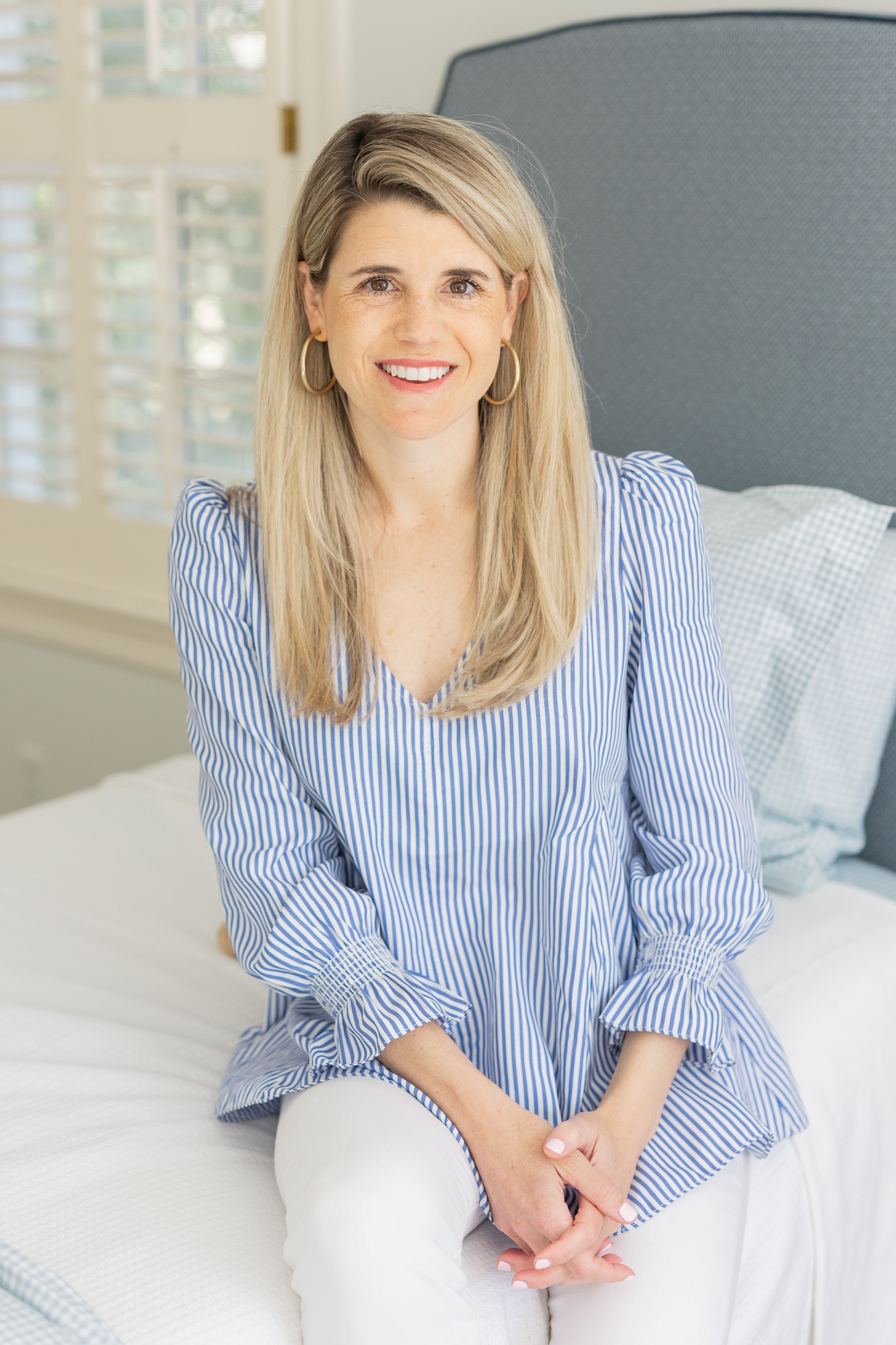 A smiling woman with blonde hair, wearing a blue and white striped blouse and white pants, sitting on a bed in a bright room with white shutters in the background.