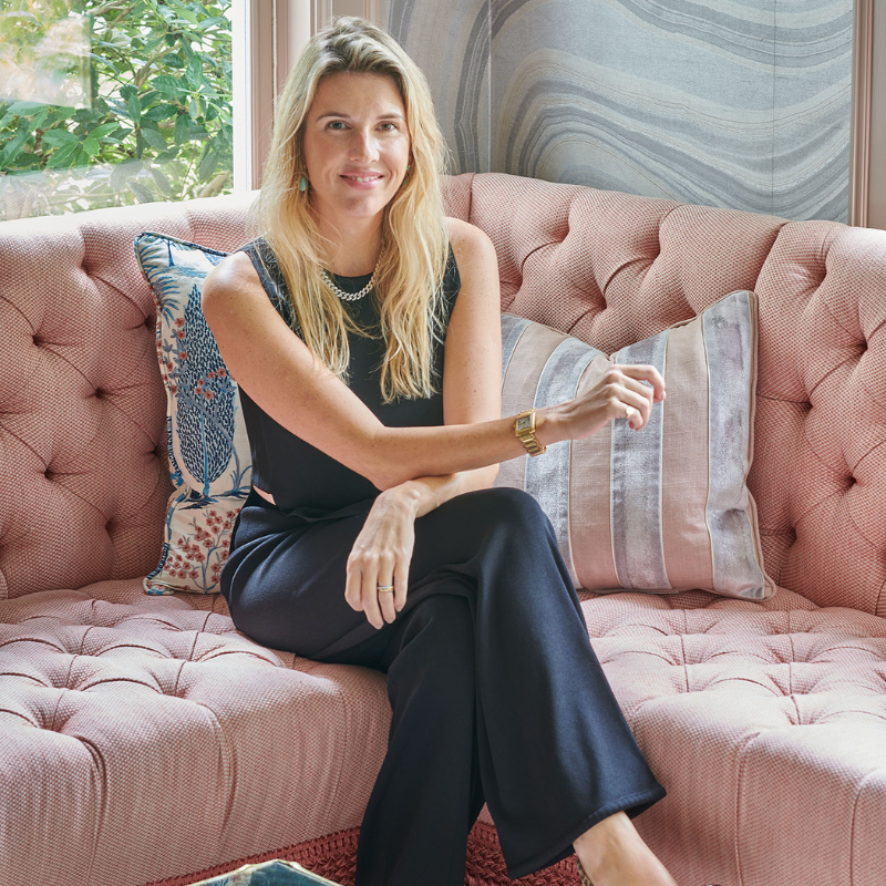 A woman with blonde hair sitting on a pink tufted sofa, smiling, wearing a black sleeveless top, black pants, and gold jewelry, in a room with a window showing greenery outside.