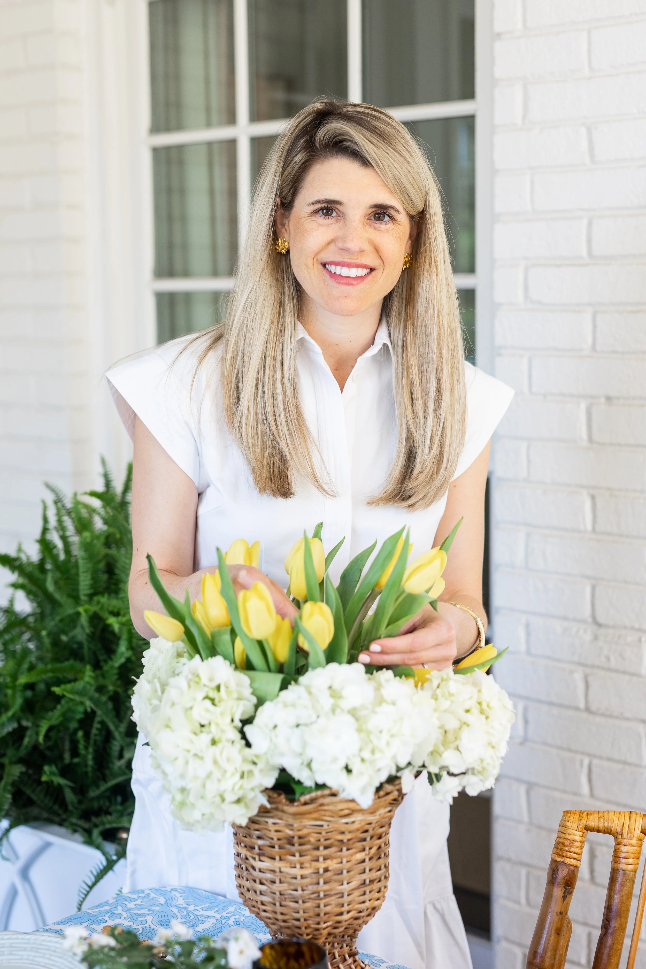A woman with blonde hair wearing a white shirt and gold earrings standing behind a basket of yellow and white flowers, smiling at the camera.