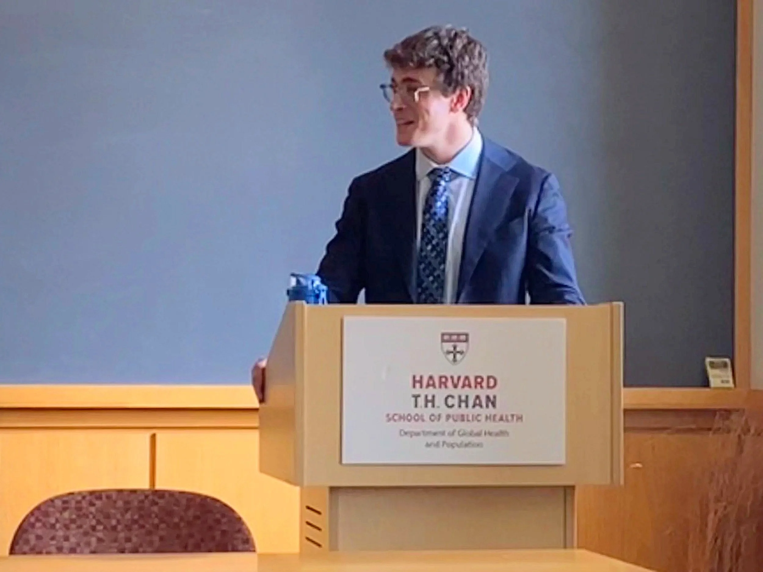 A man in a suit standing behind a podium at Harvard's School of Public Health, Department of Global Health and Population, giving a presentation or lecture.