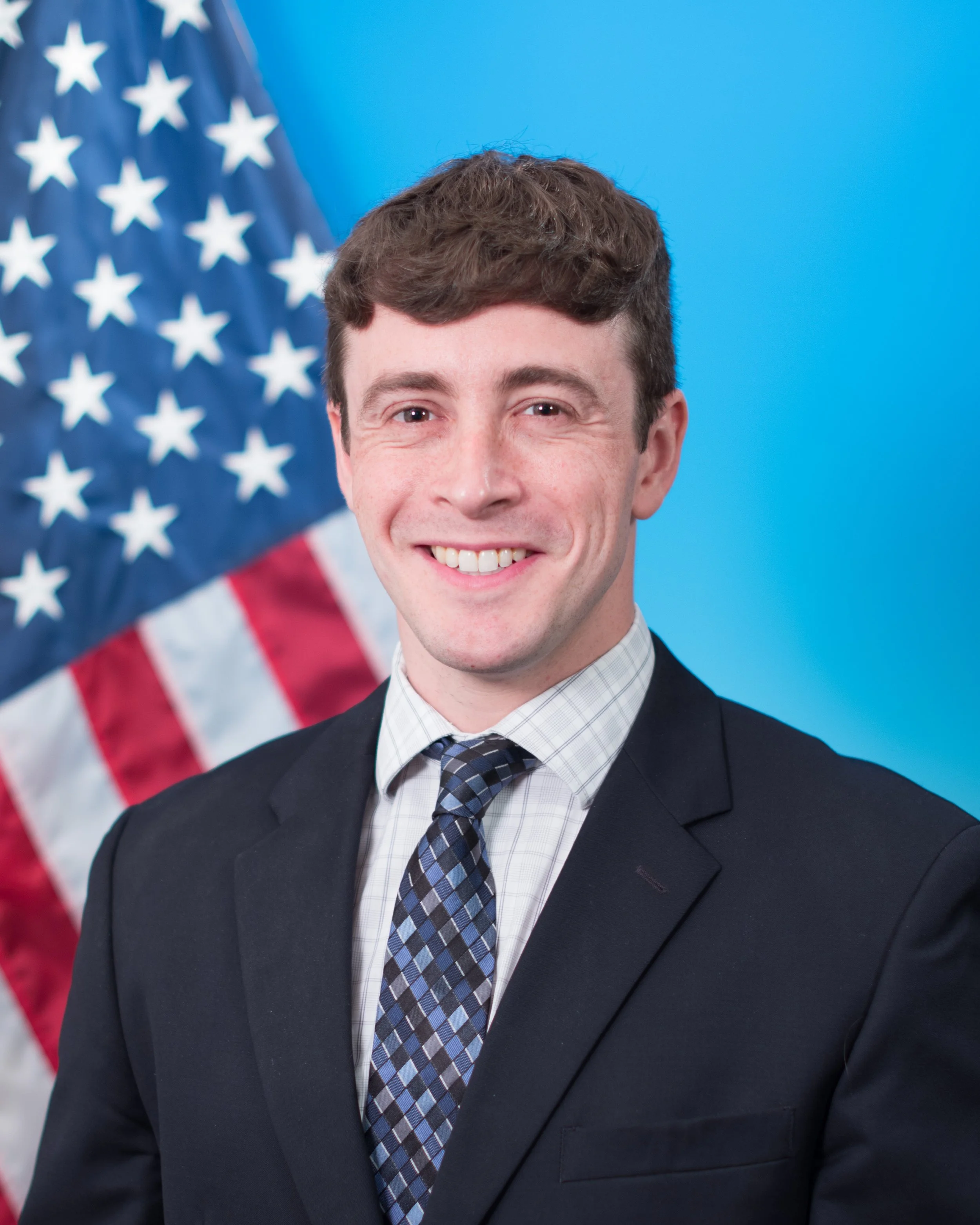 A man in a suit standing in front of an American flag with a blue background.