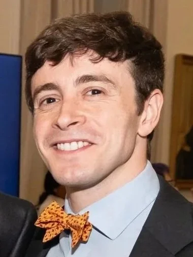 A young man with light skin and dark brown hair wearing a light blue shirt, a dark blazer, and an orange bow tie with a black pattern, smiling at the camera.