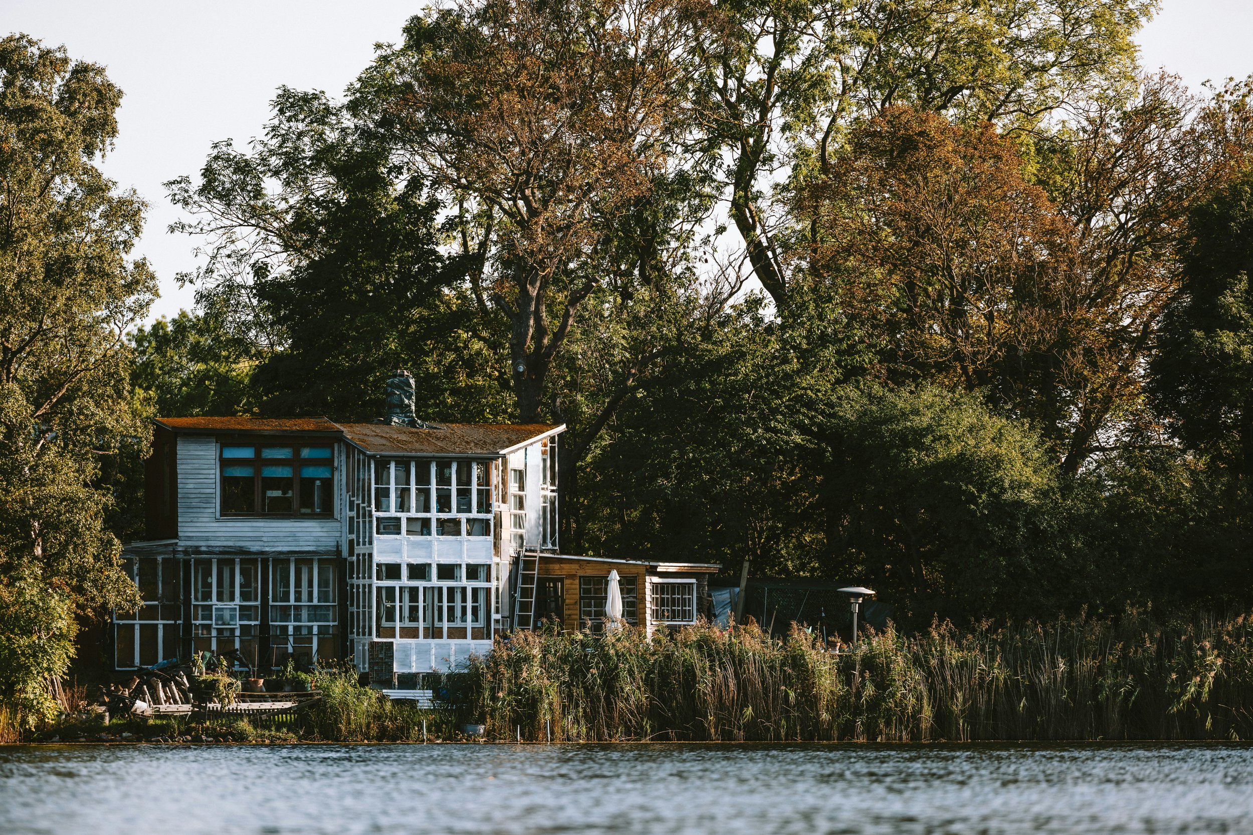 A house under construction near a body of water, surrounded by trees, with some parts of the house covered in white sheathing and reflecting the sunlight.