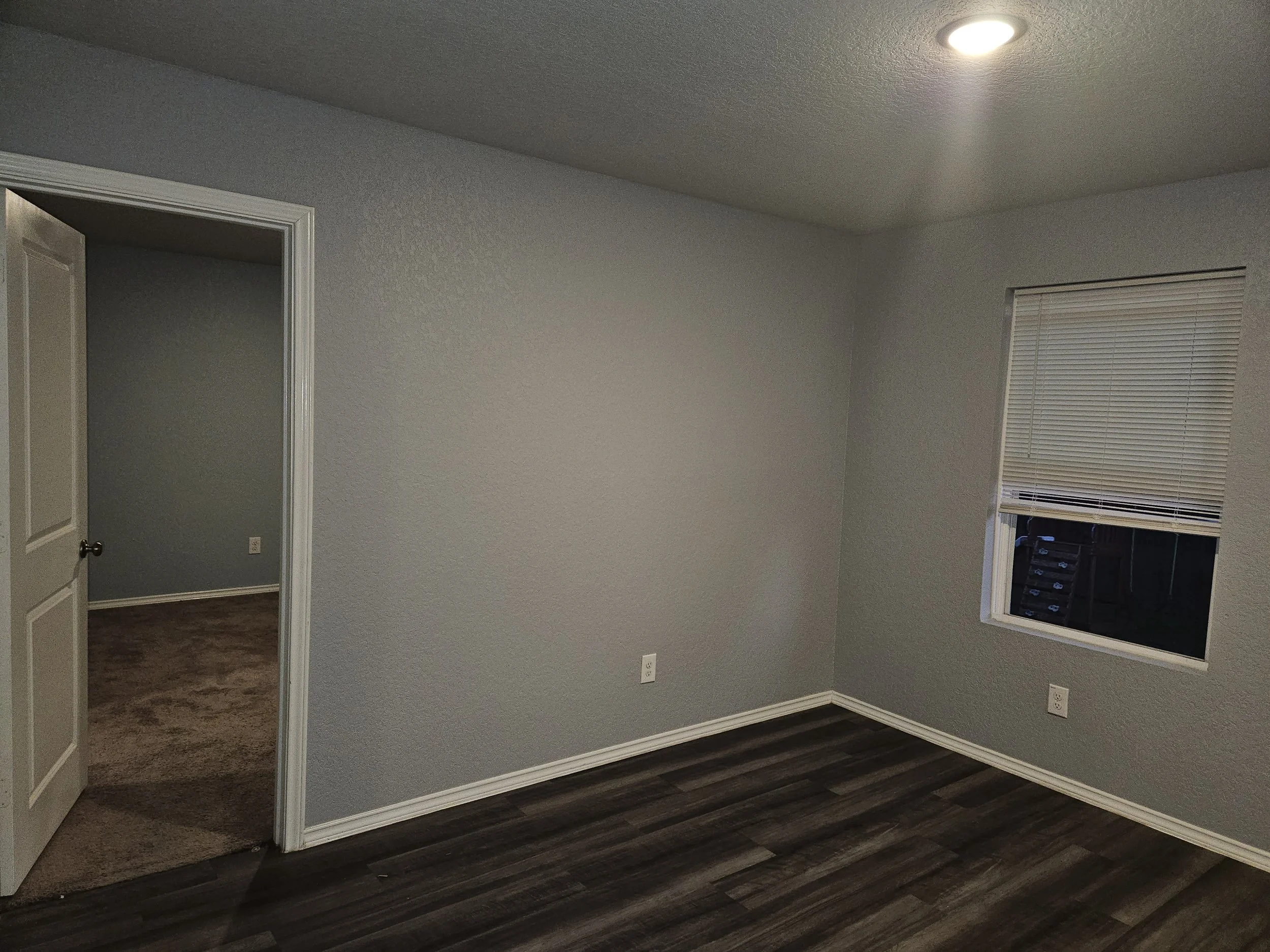 Empty room with gray walls, dark wood flooring, a window with closed blinds, and an open door leading to another room with similar gray walls and brown carpet.