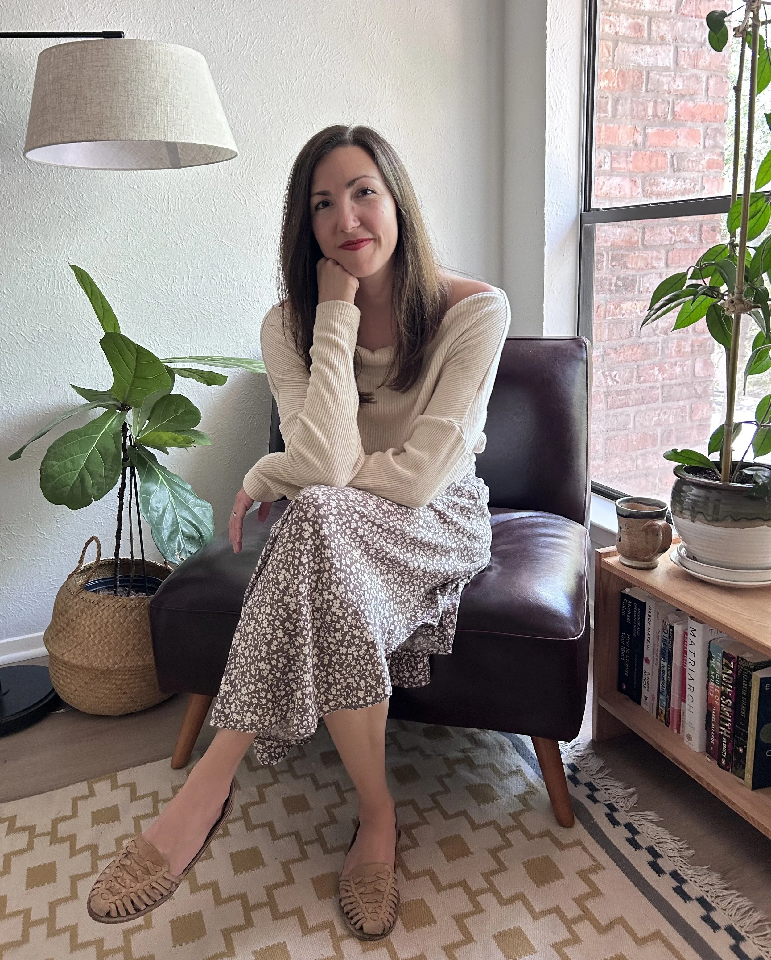 A woman sitting on a dark brown leather chair in a cozy room, surrounded by houseplants and books, with a brick wall and large window behind her.
