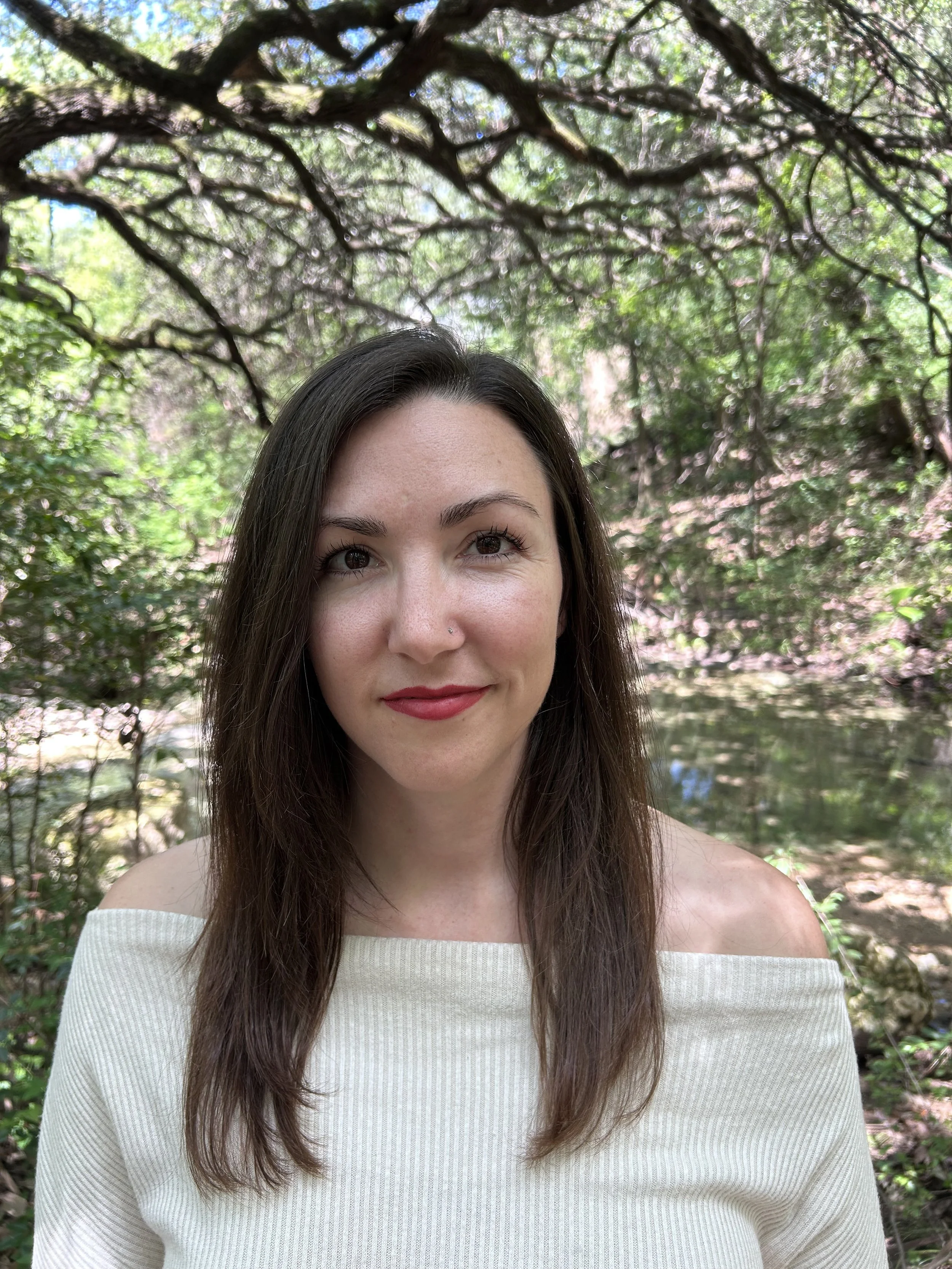 A young woman with long brown hair, wearing a white off-shoulder top, smiles softly outdoors with green trees and branches overhead.