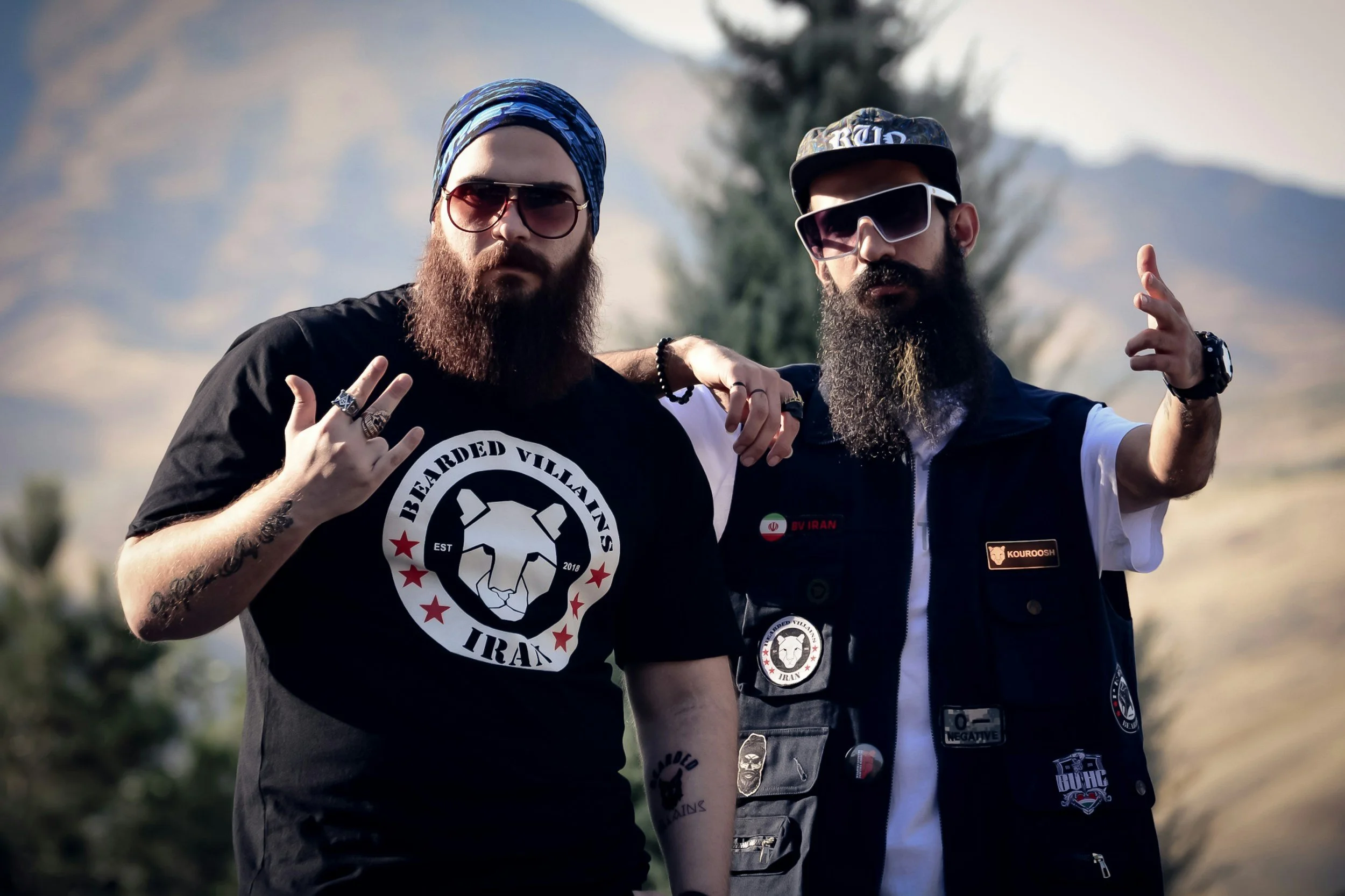 Two bearded men wearing sunglasses and casual clothing, standing outdoors with mountains in the background. One is making a gesture with his hand and the other has a hand on his friend's shoulder.