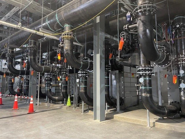 Industrial piping system with large black pipes, valves, and gauges in a facility under construction or maintenance, with orange safety cones on a concrete floor.