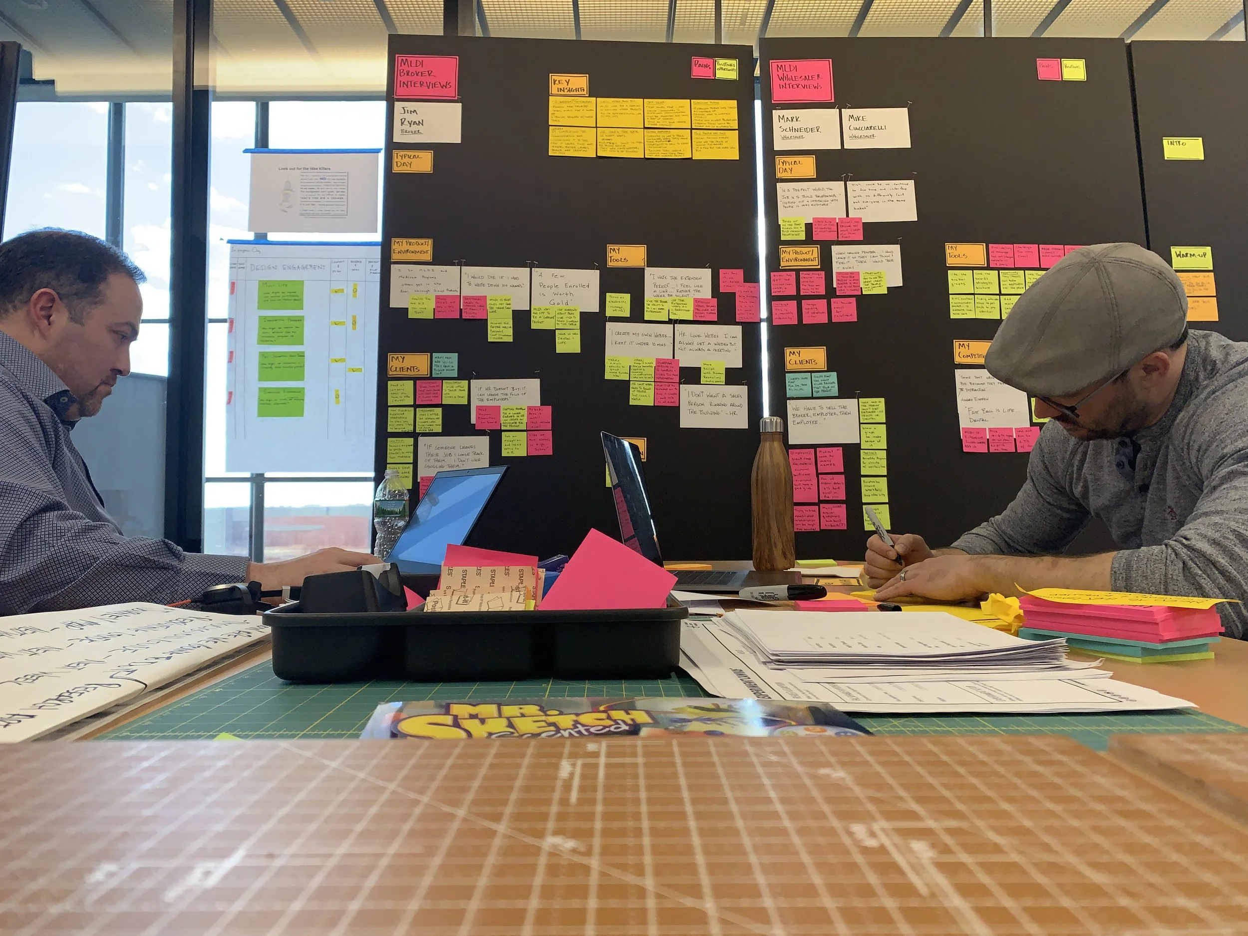 Two people working at a desk with a large black board filled with colorful sticky notes behind them. The desk has papers, notebooks, a water bottle, and laptops. The background shows windows with a city view.