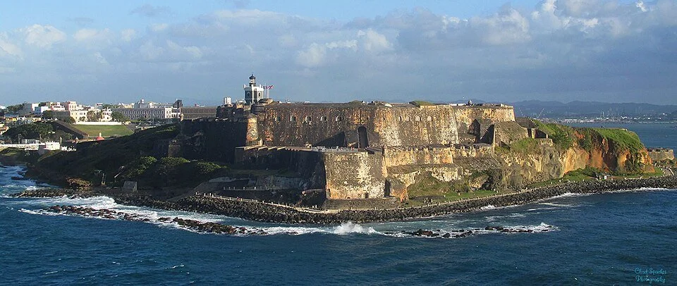 Fuerte en una isla vista desde el océano con ciudad en el fondo y cielo con nubes.