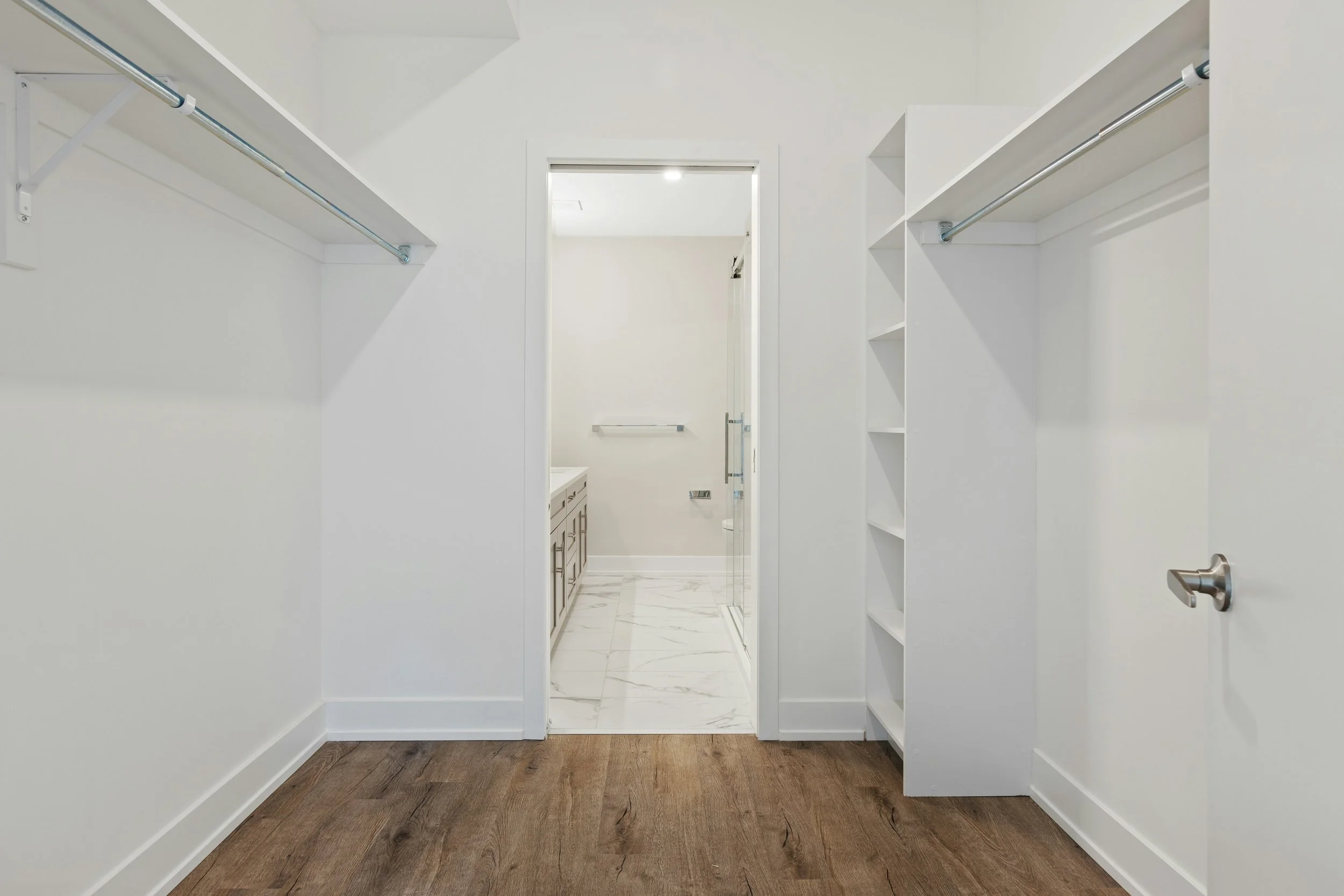 Empty walk-in closet with white walls and wooden flooring, shelves on the right, a hanging rod on the left, and a view into a bathroom with marble floor tiles, a vanity, and a glass shower door.