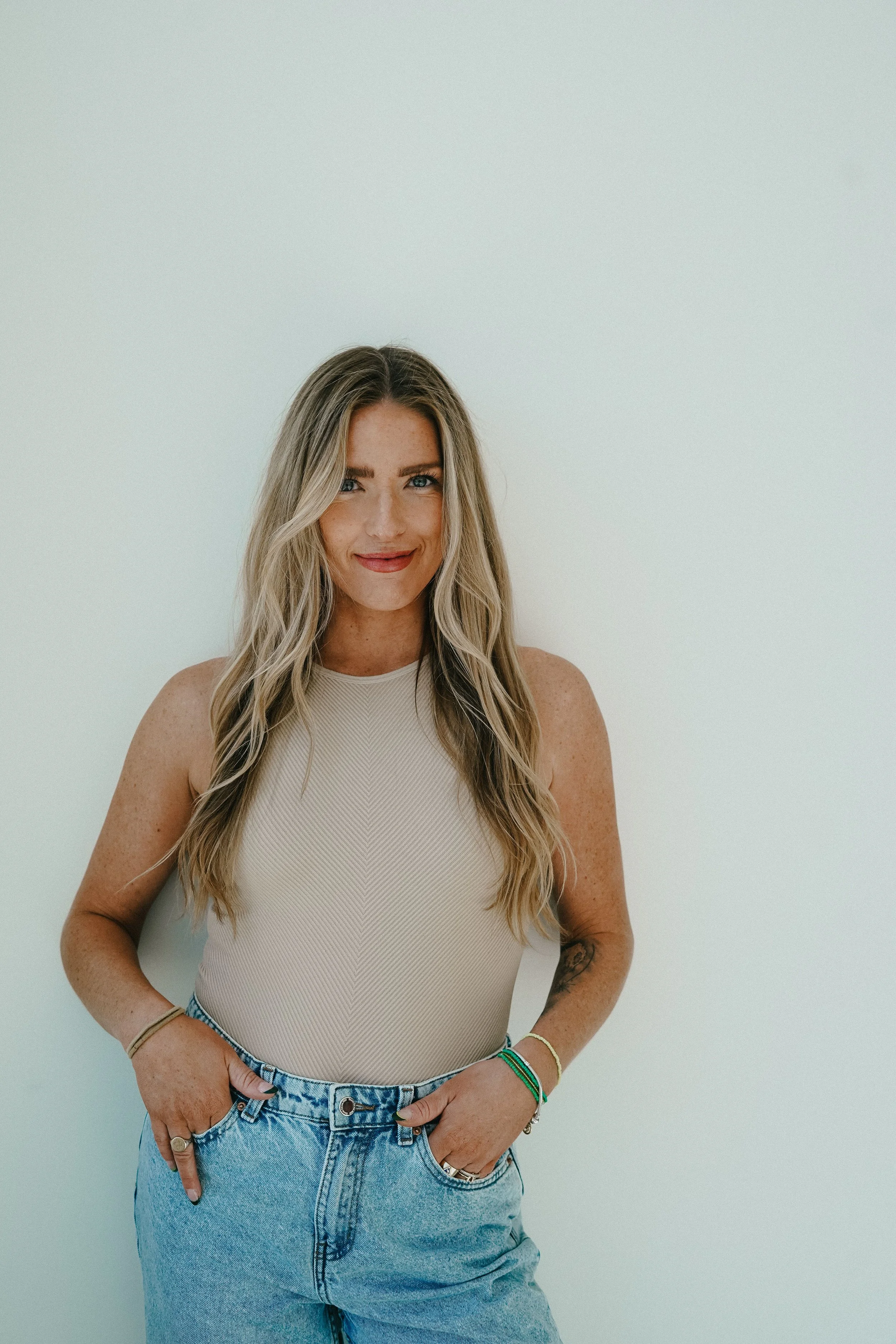 A young woman with long wavy blonde hair, wearing a sleeveless beige top and light blue high-waisted jeans, standing against a plain white wall, smiling softly with her hands in her pockets.