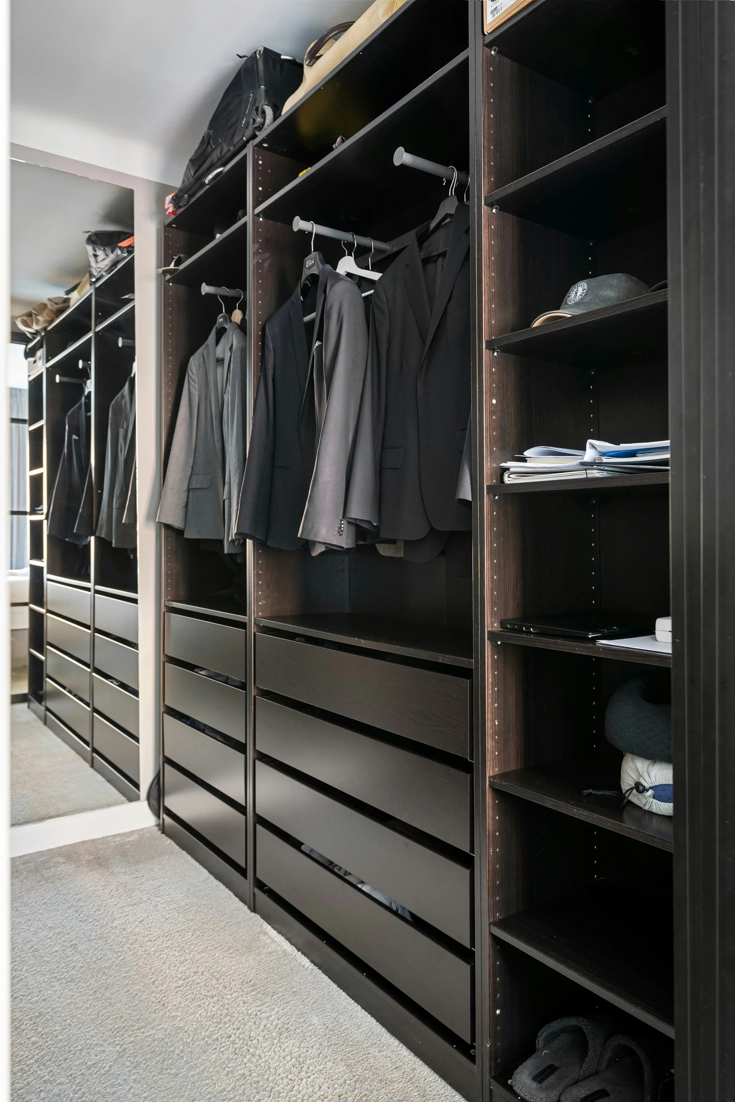 A walk-in closet with black and brown open shelving units, displaying hanging jackets and shirts, folded clothes, books, hats, and shoes.