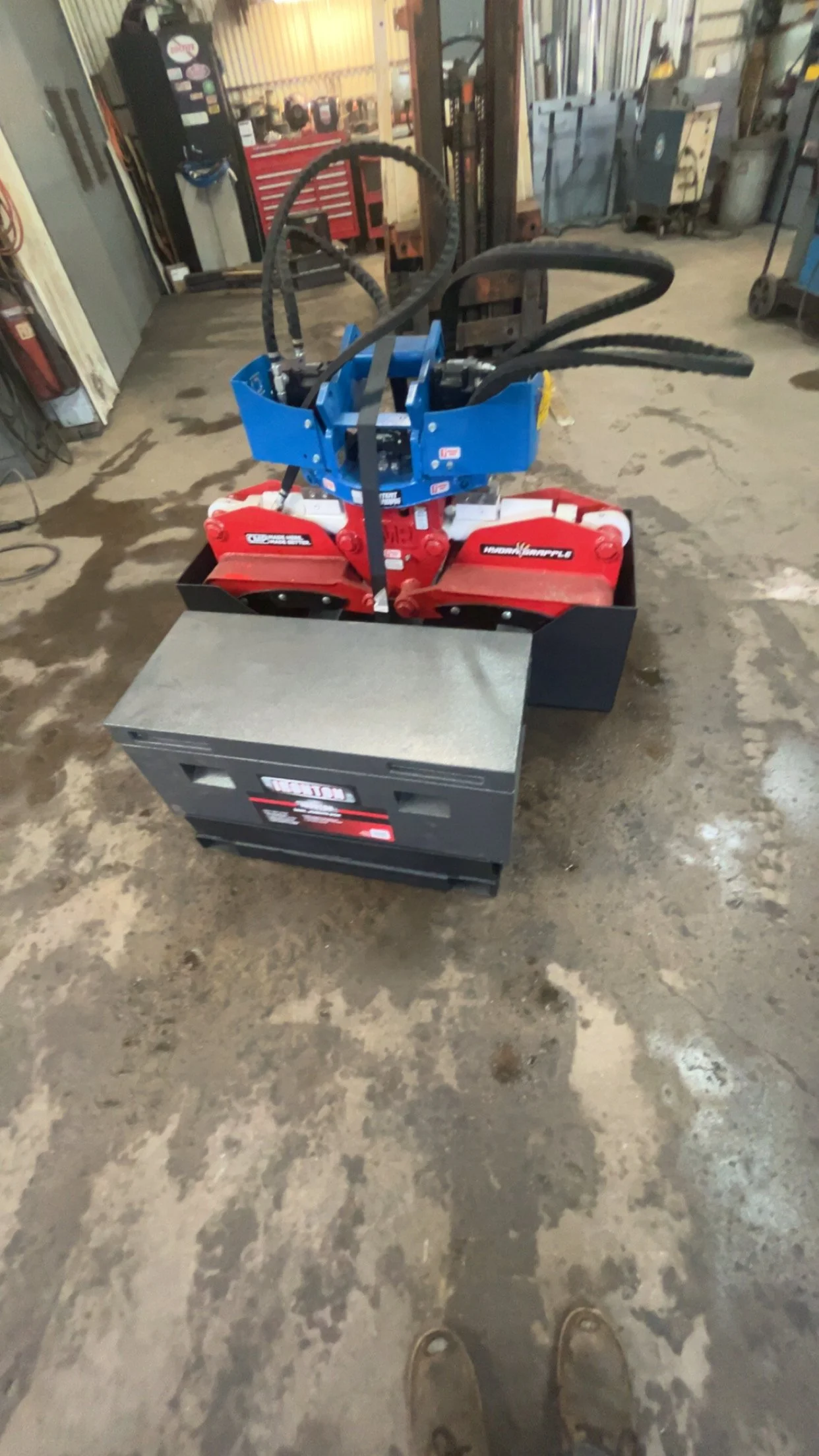 A piece of machinery with black hoses, a blue top, and red and black components is positioned on a concrete floor in a workshop or garage setting.
