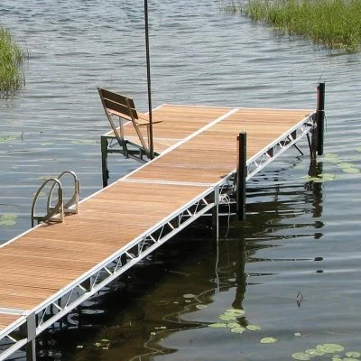 A wooden dock with a ladder extends into a lake, with a bench at the end of the dock and a flagpole.
