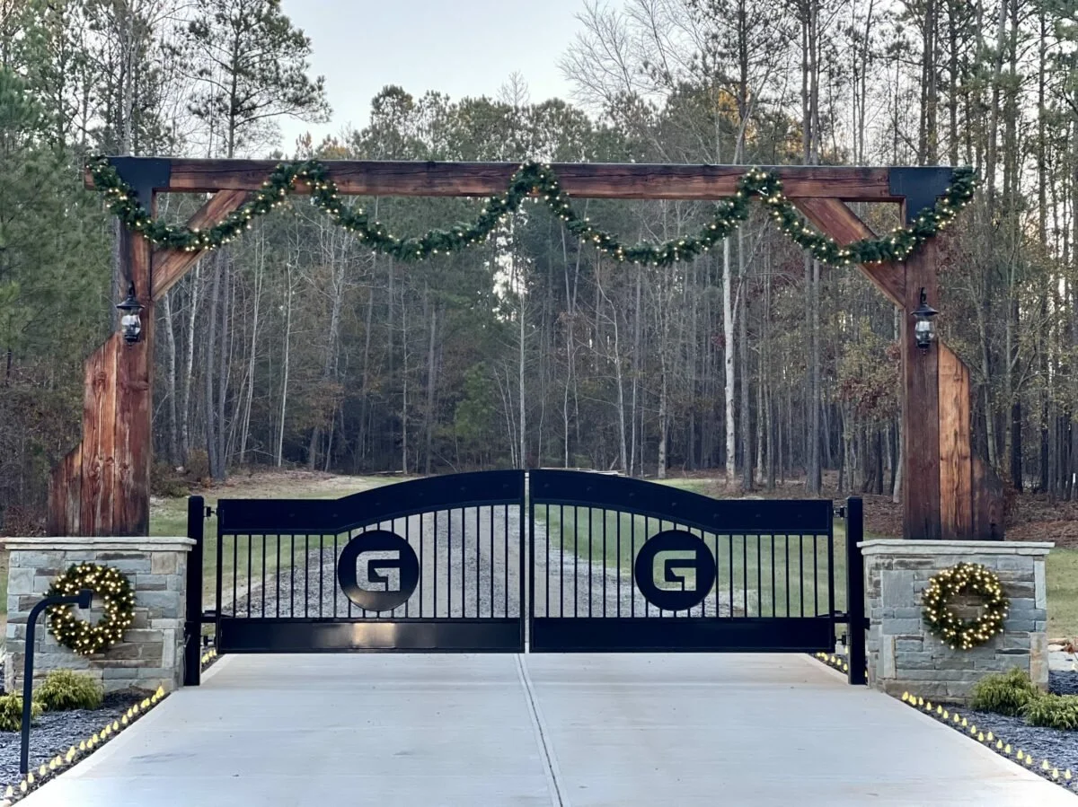 Decorated driveway entrance with a black gate, stone pillars, and a wooden arch with holiday wreaths and garlands in a wooded area.
