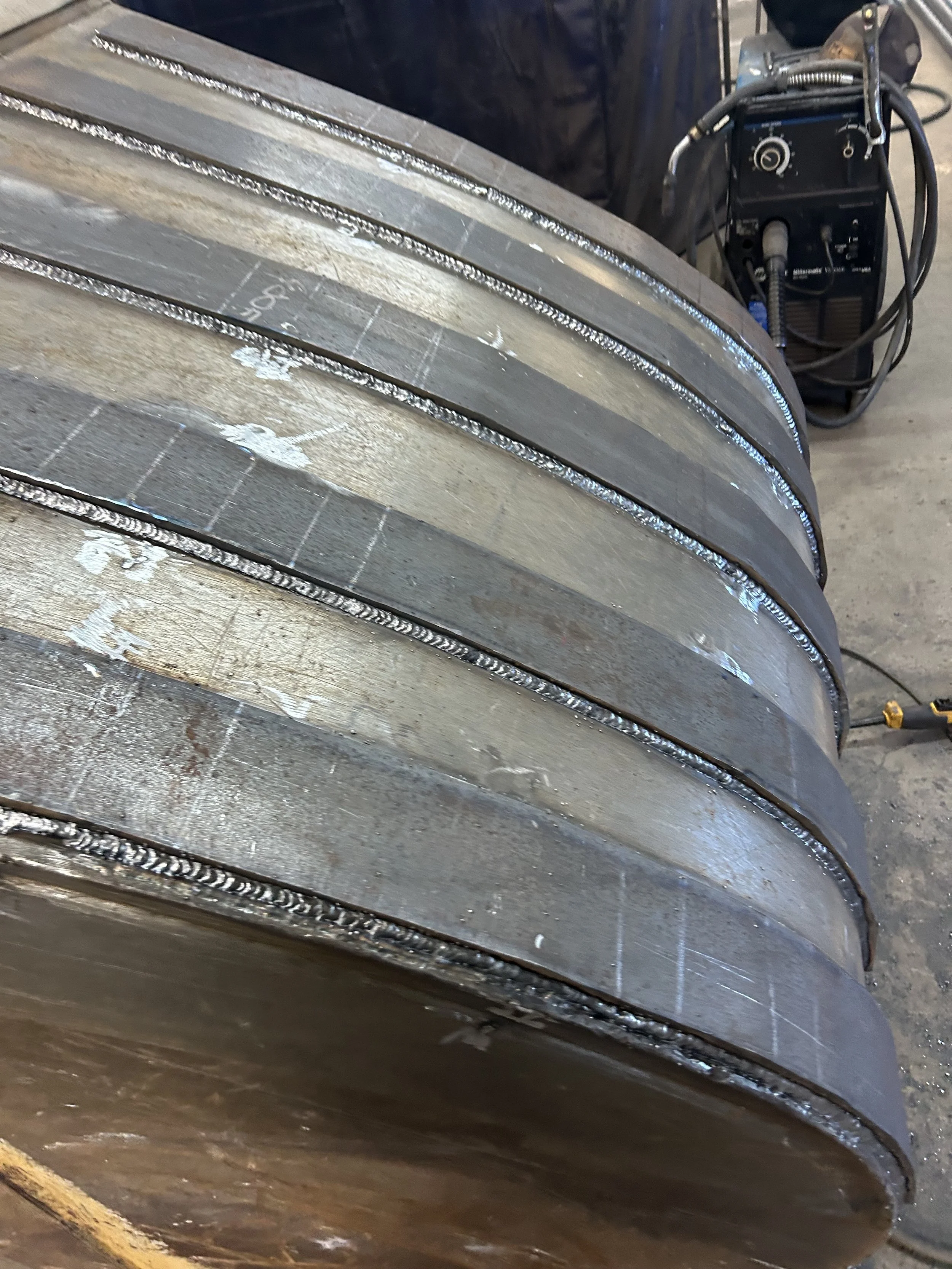 Close-up view of a metal welding process on large, curved metallic components with a welding machine in the background.