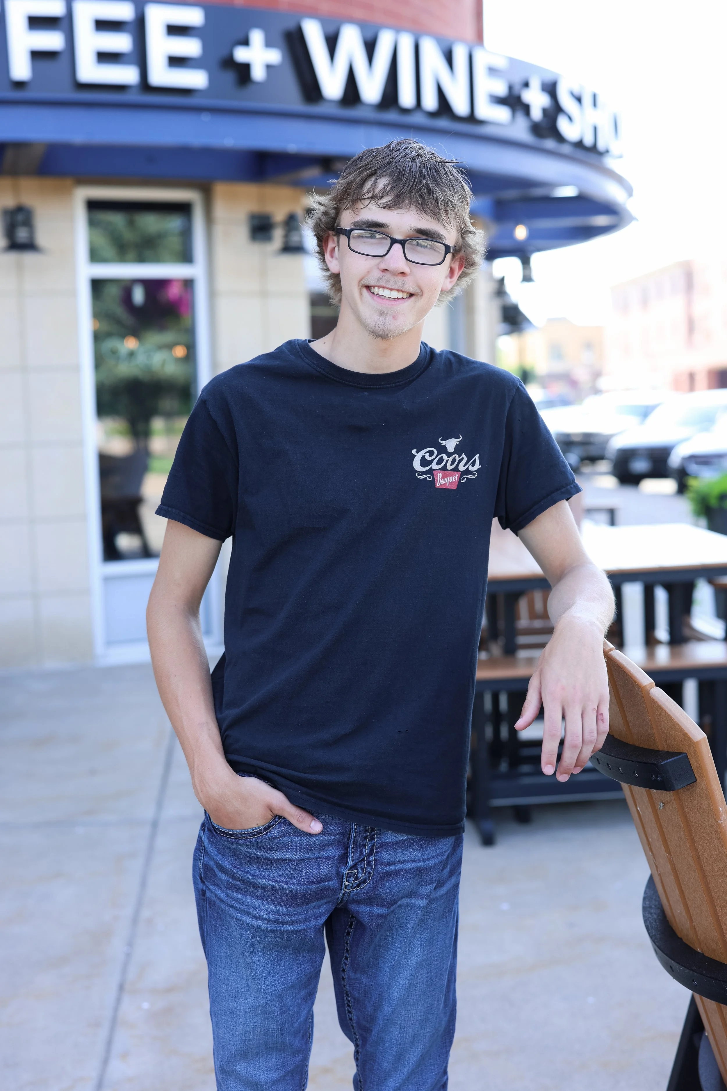 A young man with glasses and messy hair smiling while standing outside a restaurant with a sign that says "Coffee + Wine + Sushi." He is wearing a black t-shirt with a Coors logo and jeans, leaning with his left hand on a wooden chair.