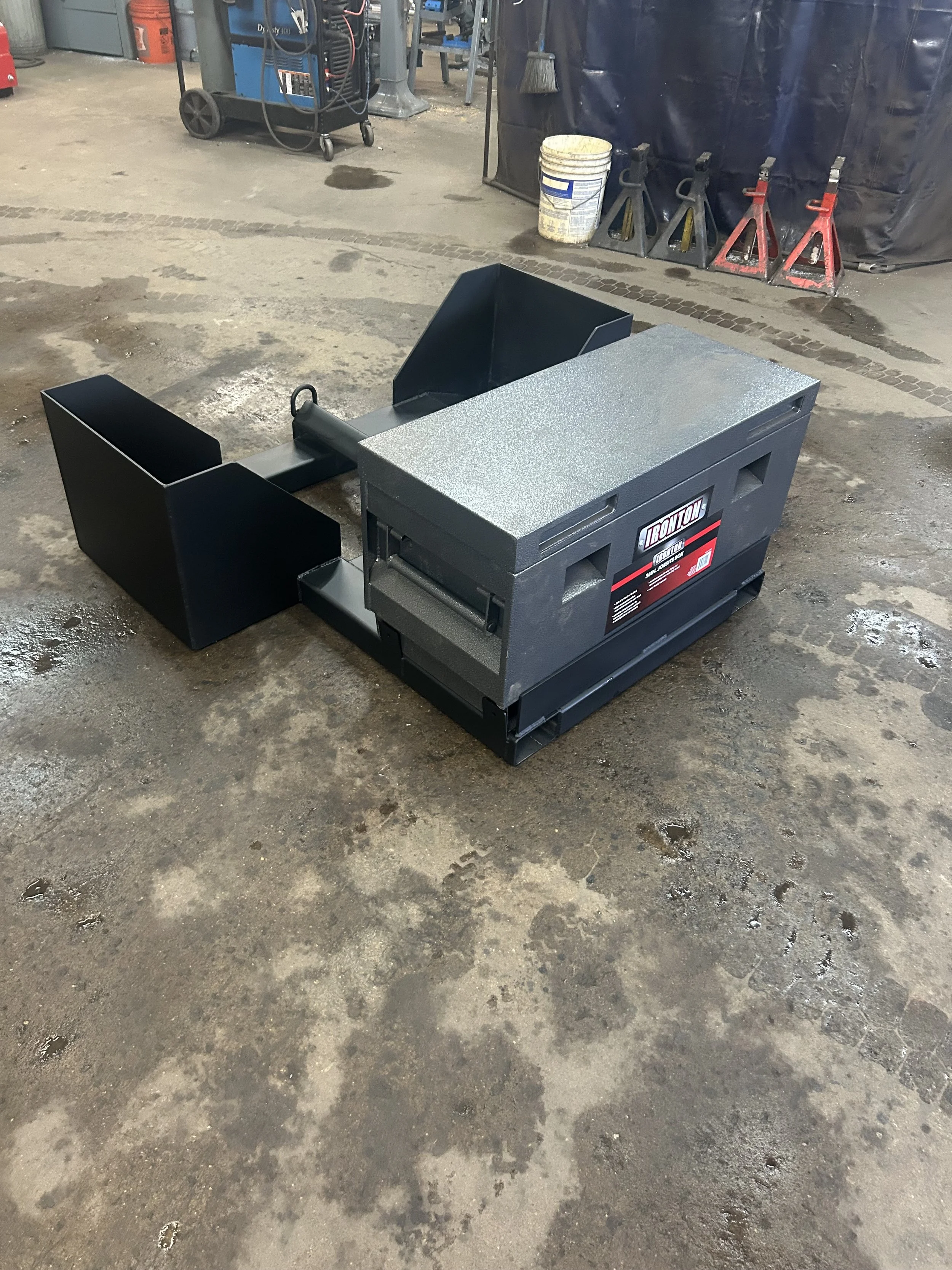 A black, metallic, portable welding table with a textured surface, placed on a worn concrete workshop floor. In the background are work tools and equipment including a bucket, jack stands, and a welding machine.