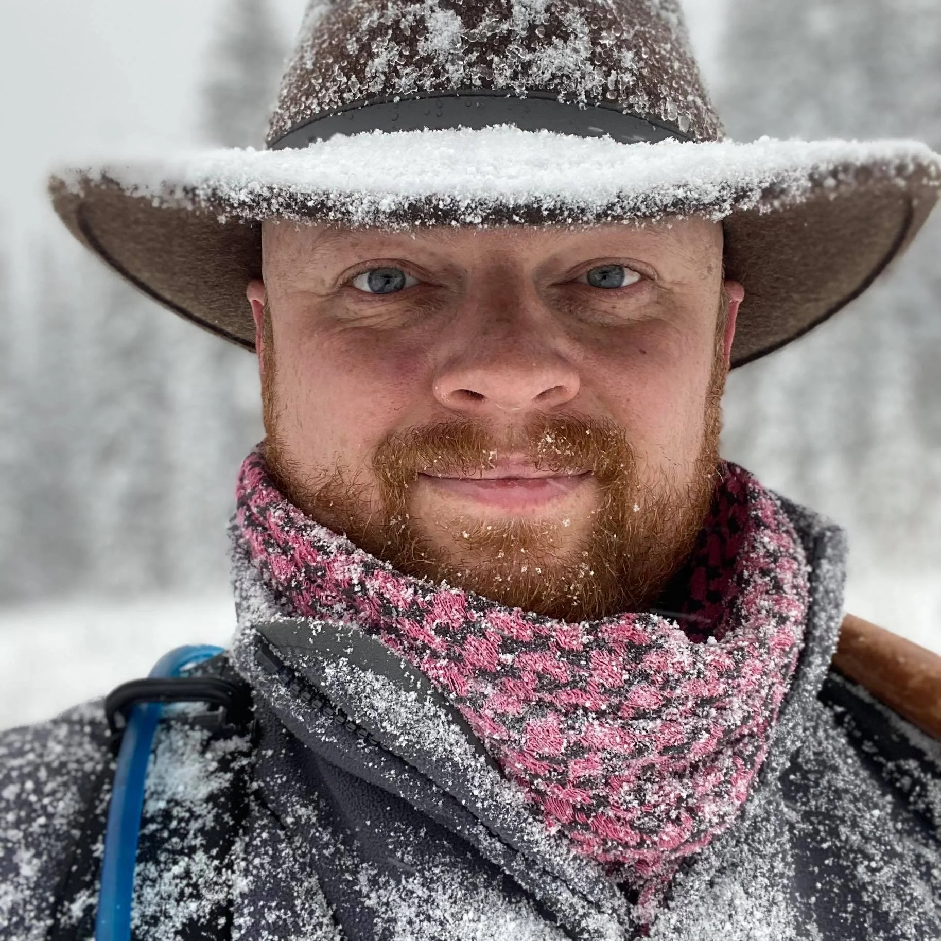 Close-up image of a man's face partially covered with snow. He is wearing a large wide-brimmed hat, with snow on it, and a colorful scarf around his neck. The background is blurred snow.