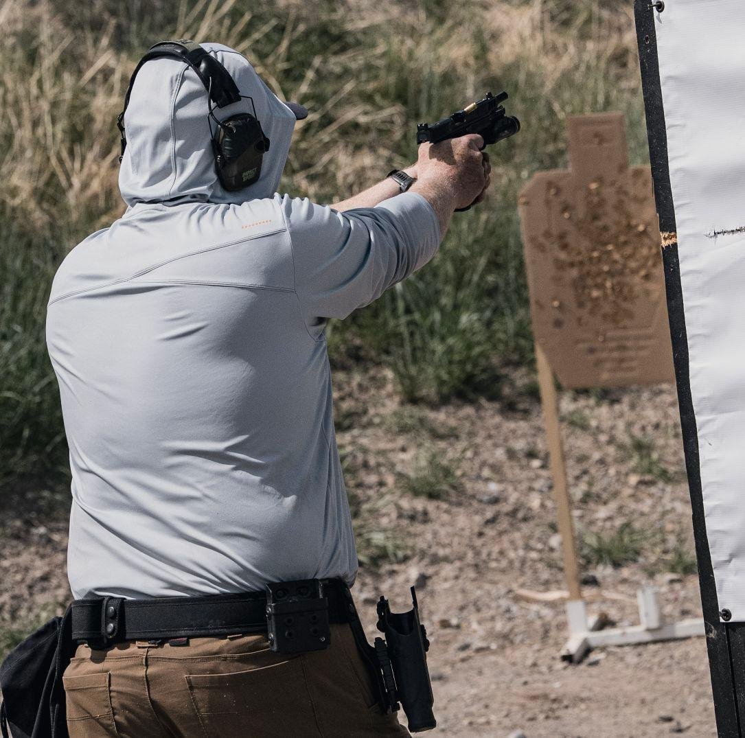 Person wearing a gray hoodie and khaki pants aiming a handgun at a shooting target outdoors, with hearing protection on.