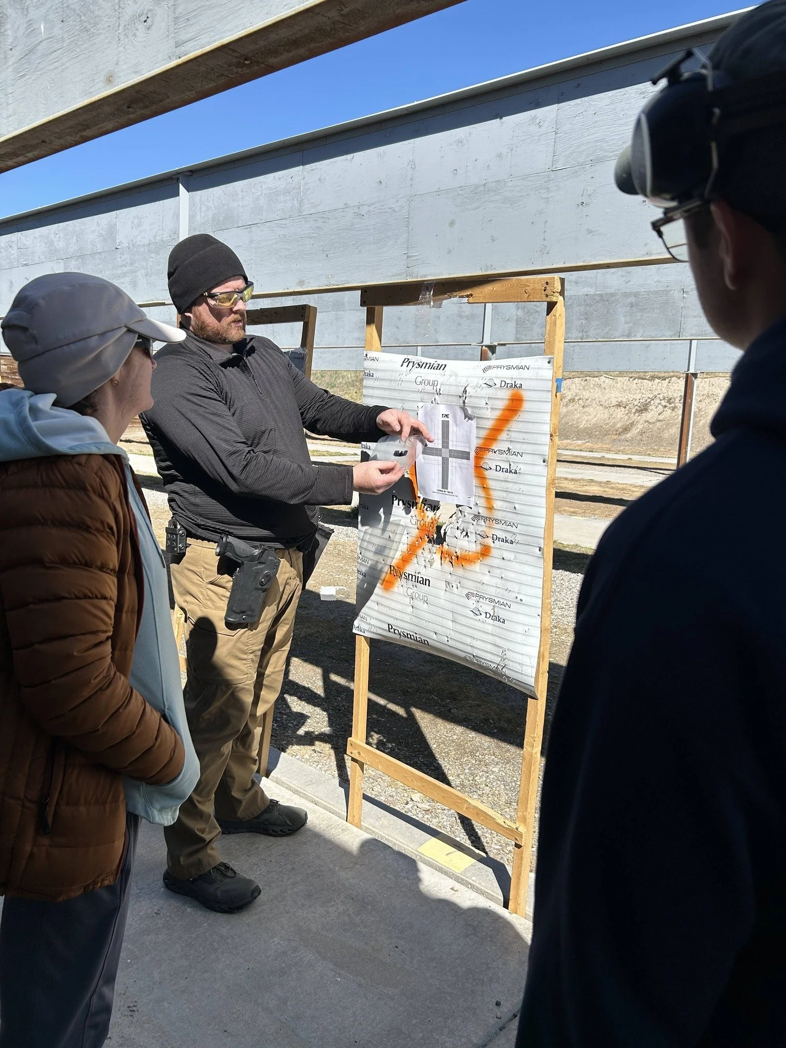 A man explaining shooting data with three people listening, at an outdoor shooting range, showing a target station with a sheet of paper and spray-painted lines.