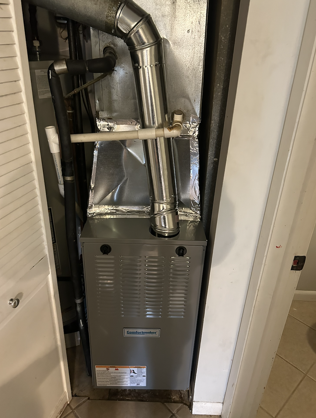 A furnace unit in a closet with metallic pipes and ducting connected to it.