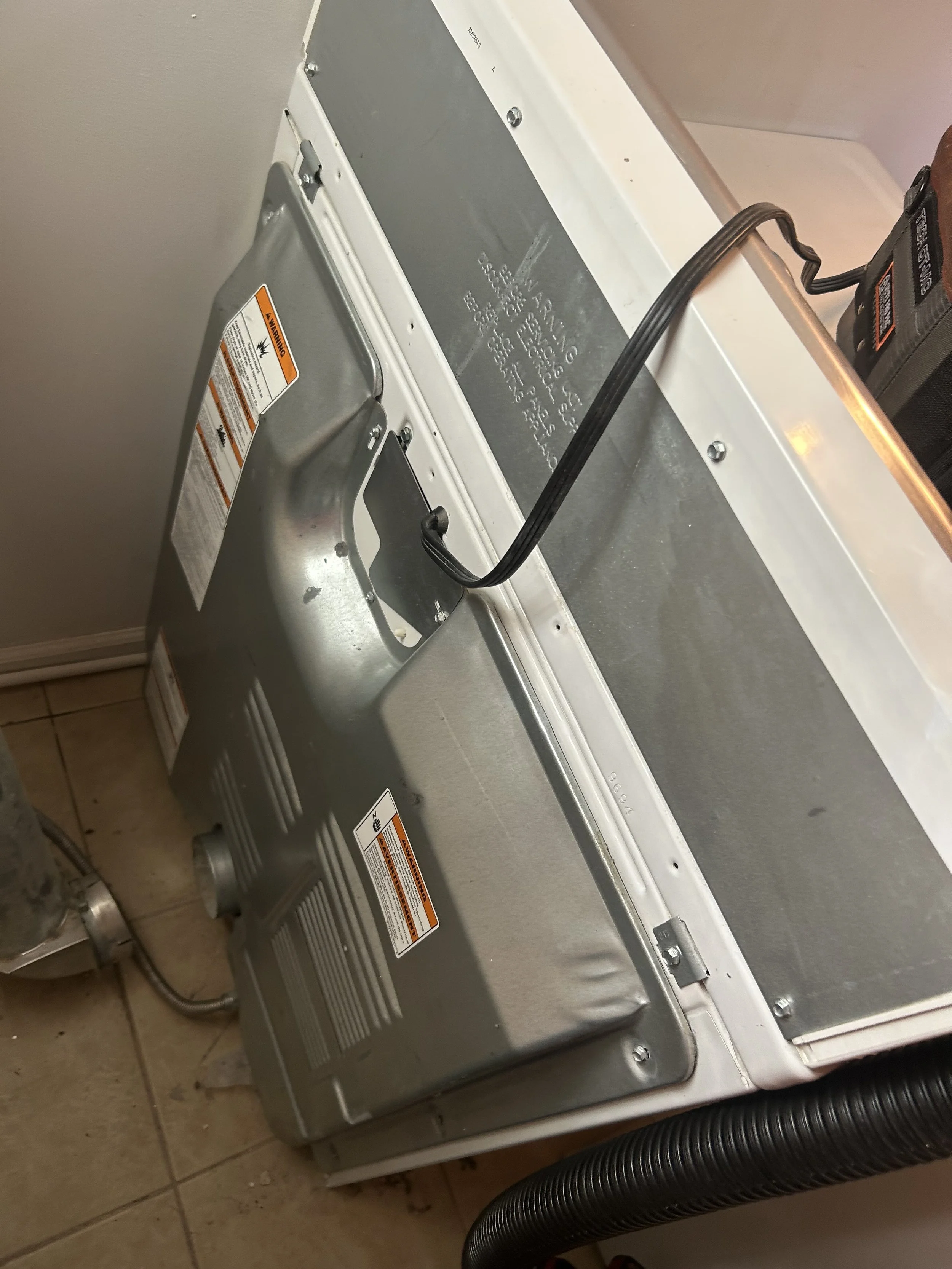 Image of a washing machine or dryer with white and metallic parts, electrical cord, and warning labels, placed in a laundry area with tiled floor.