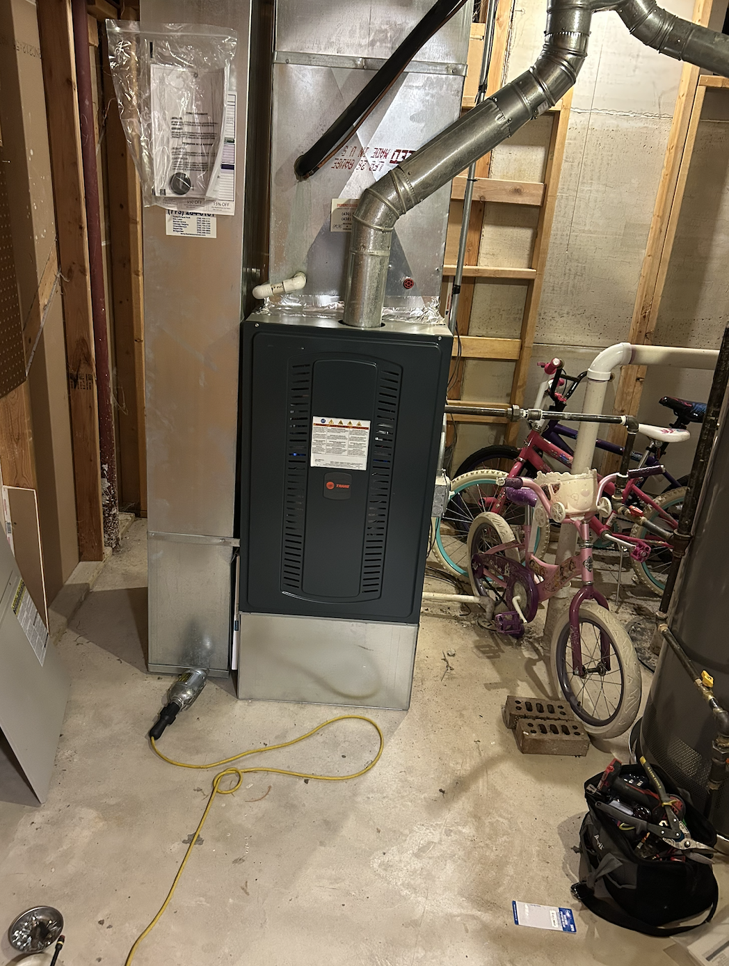 A furnace or boiler installed in a basement, with a duct pipe connected on top, a red switch on the front, and a yellow extension cord on the concrete floor. To the right, there are two children's bicycles, one pink and one purple, leaning against th