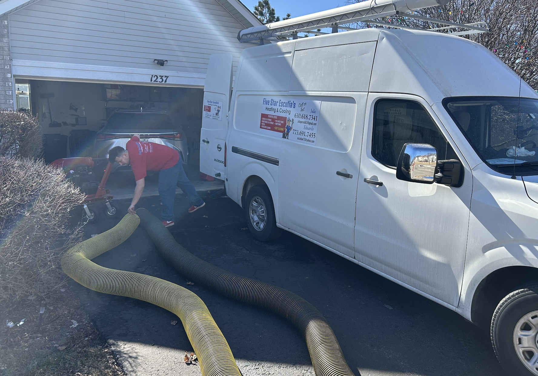 A worker wearing a red shirt and dark pants is using a vacuum hose to clean air ducts, a white service van with signage. The service van is labeled for Five Star Esucursal's Heating & Cooling, parked in front of a house with a partially open garage.