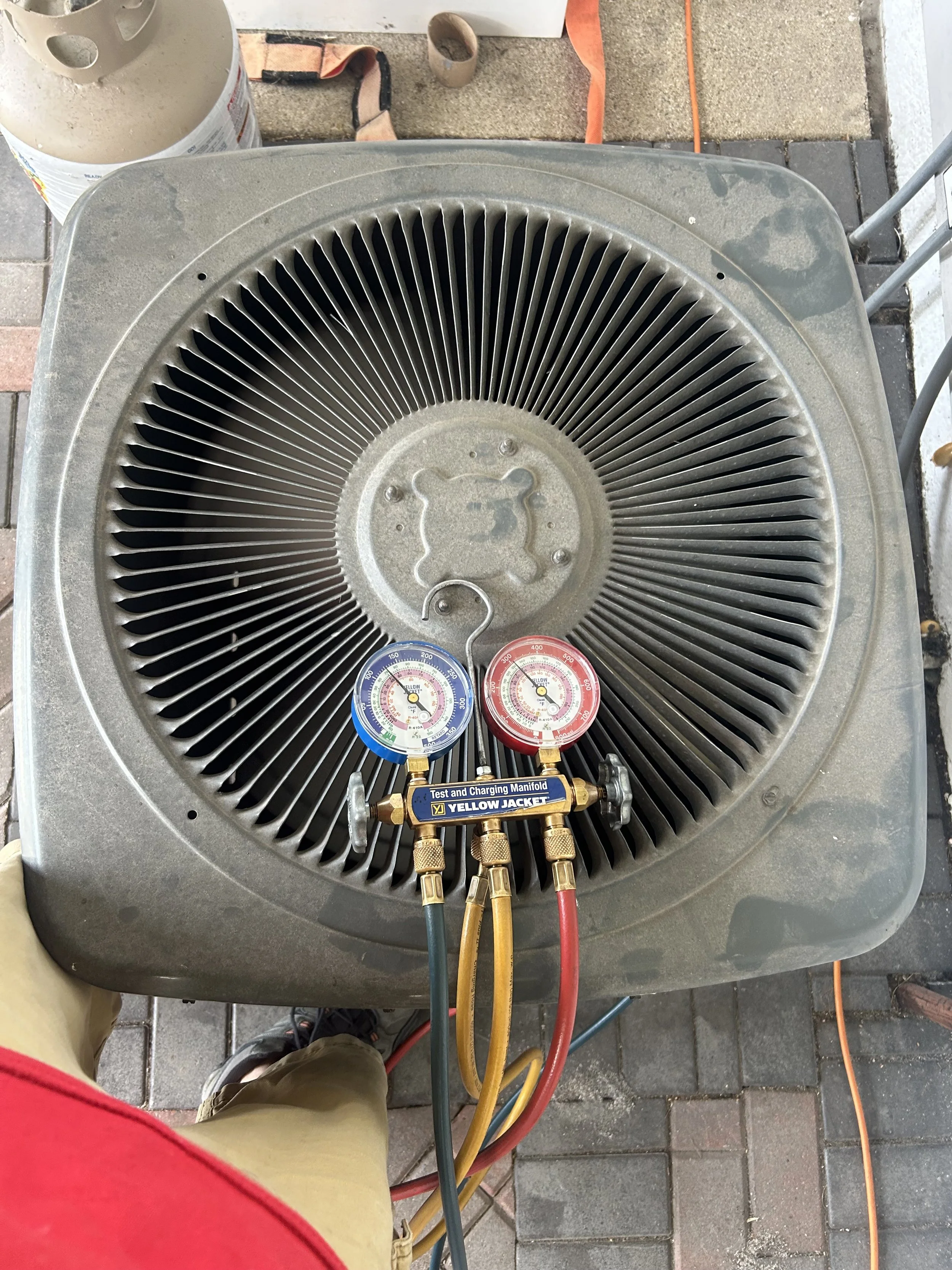 Top-down view of an HVAC air conditioning unit with gauges attached, located outdoors on a tiled surface, with some tools and equipment nearby.