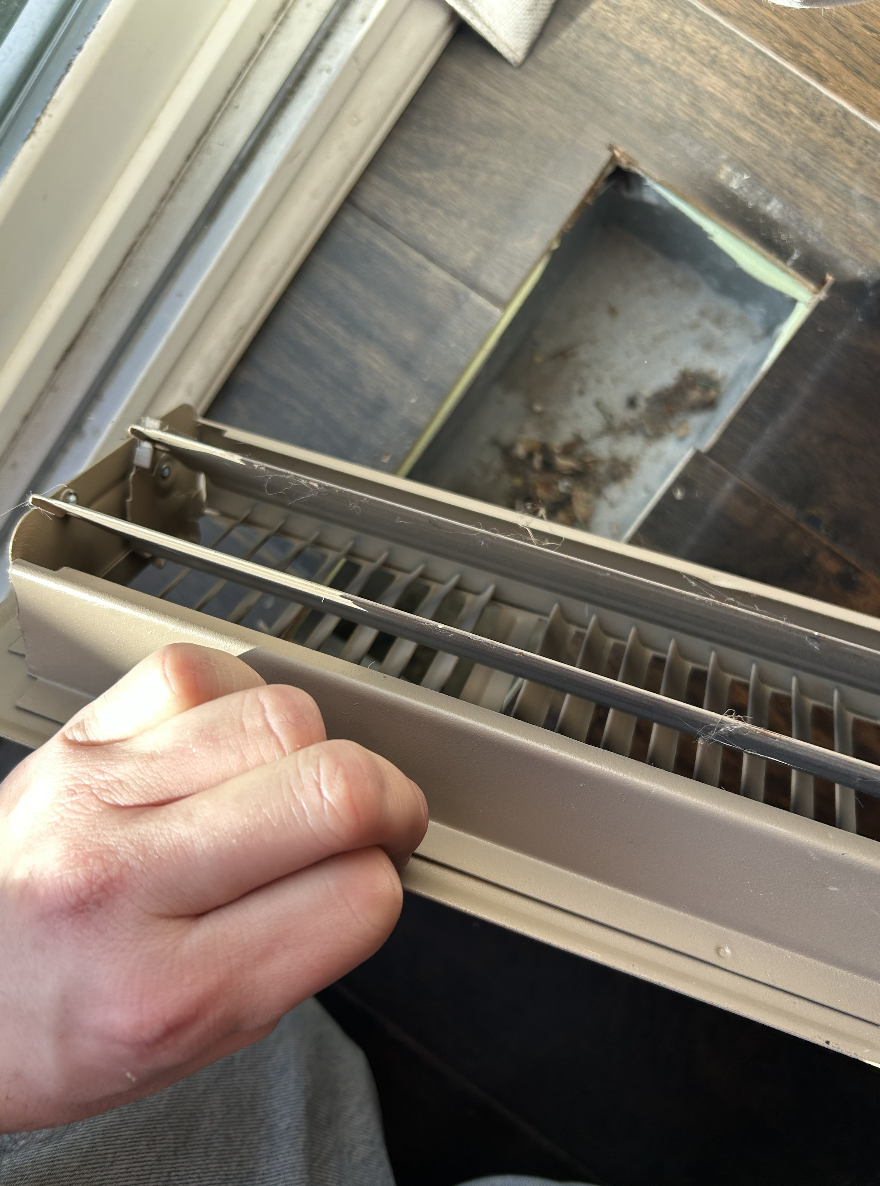 A person holding the cover of an air vent or HVAC grille, revealing dirt and debris inside, on a hardwood floor near a wall.