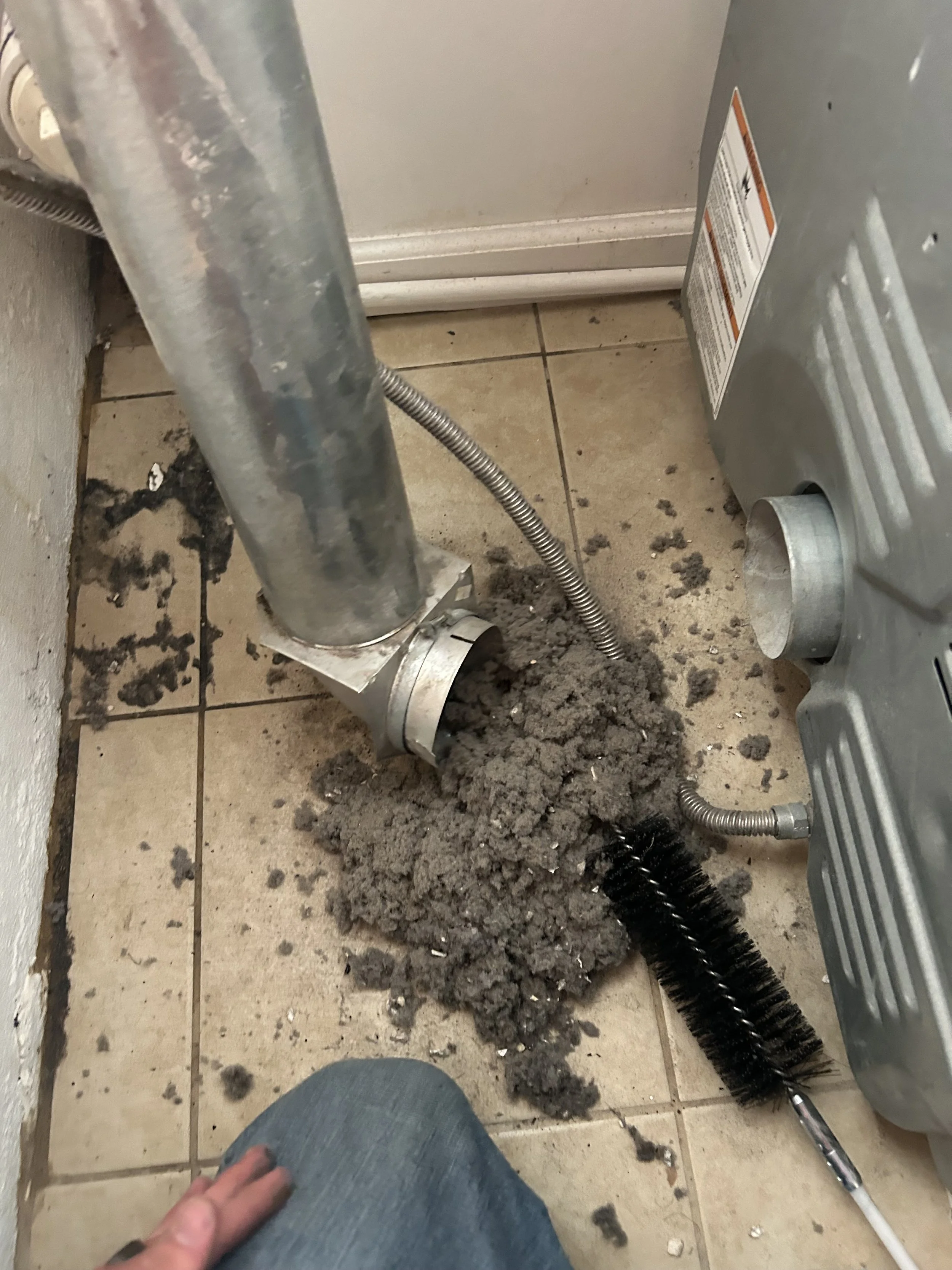 A plumber's workspace showing a drain pipe disconnected from the wall, with dirt and debris around it on the tiled floor. A person wearing jeans is partially visible in the lower left corner.