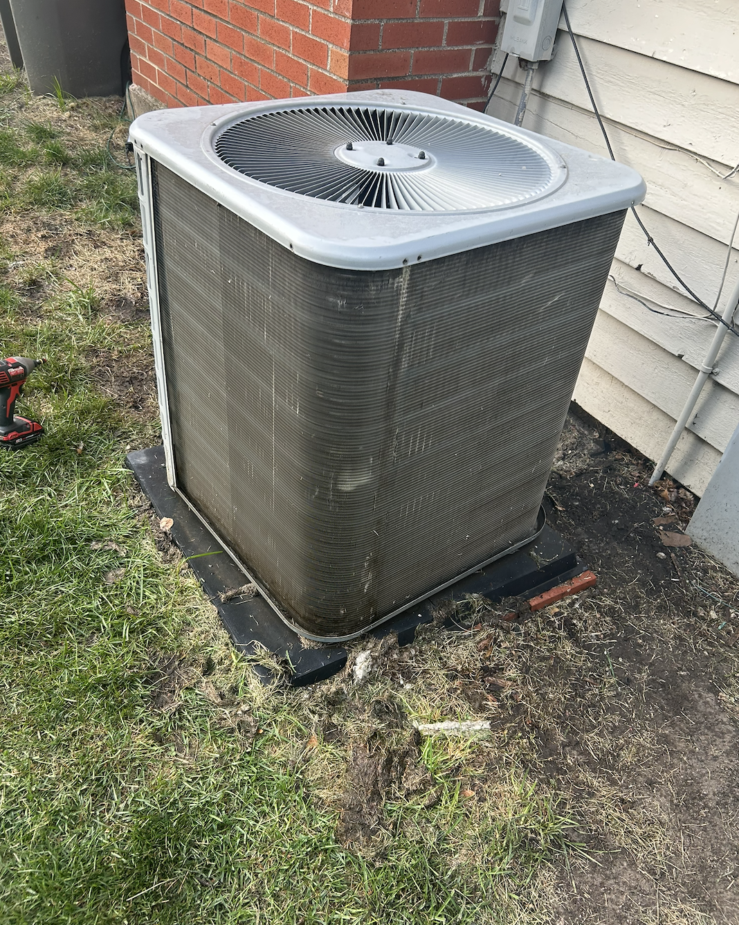 Outdoor air conditioning unit beside a brick and wooden house wall, with some grass and dirt ground, and a power cord and tool nearby.