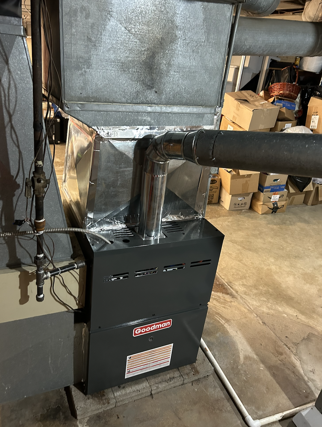 A Goodman HVAC furnace unit connected to ductwork with a black pipe and silver vent pipe.