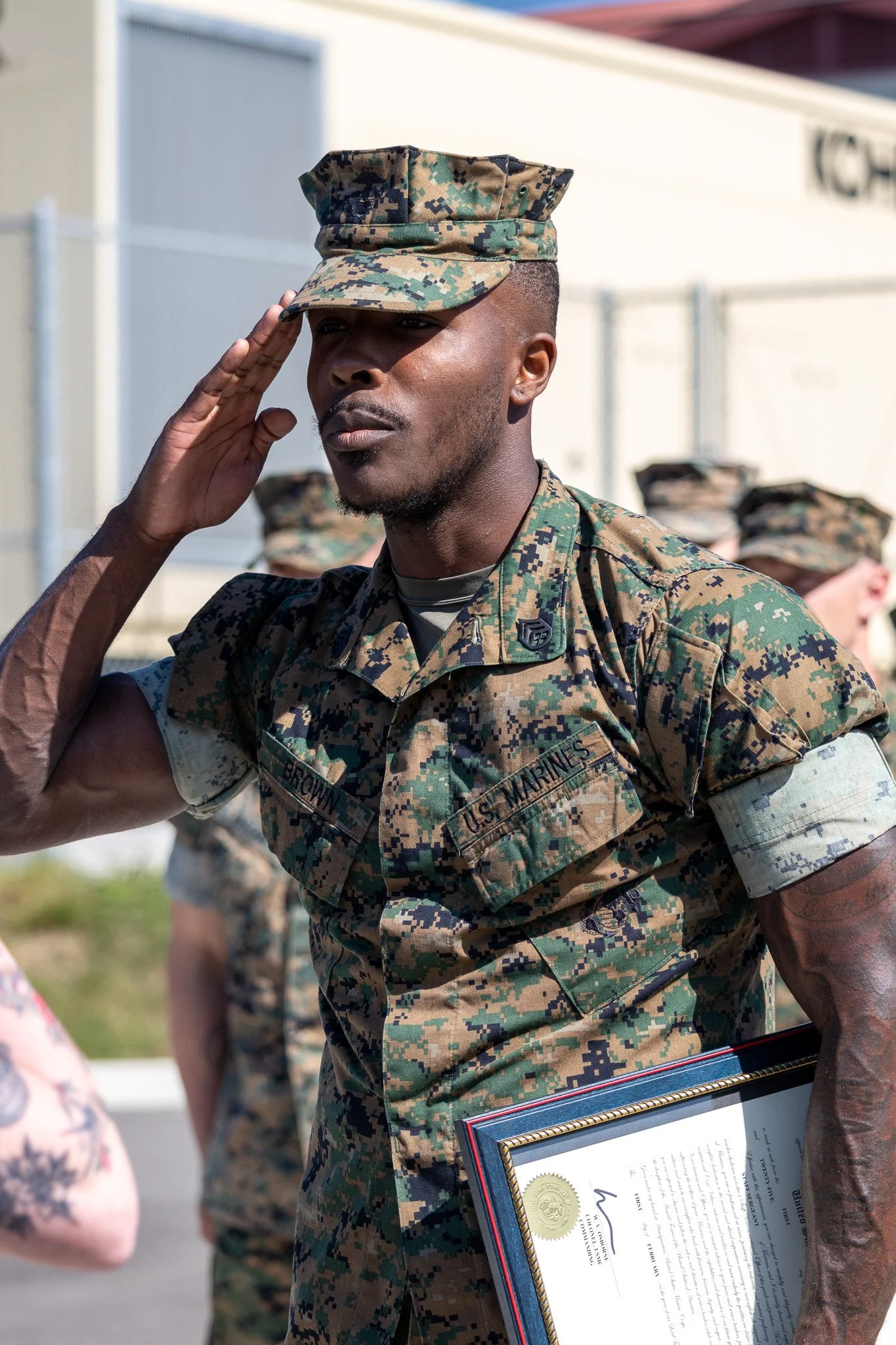 A man in a U.S. Marine Corps uniform salutes while holding a framed certificate, with other Marines in the background.
