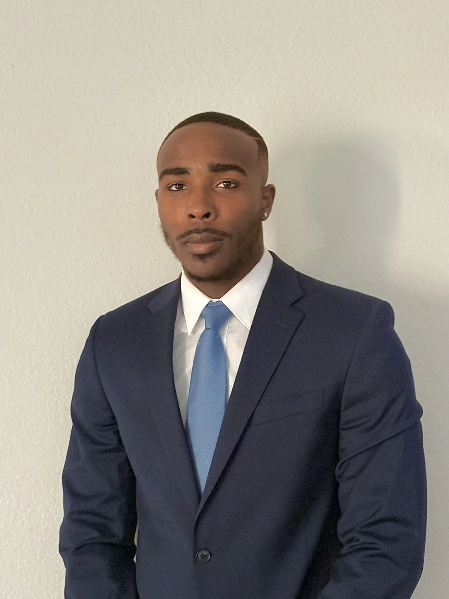 A young man wearing a dark navy suit with a white shirt and light blue tie, standing against a plain light-colored wall.