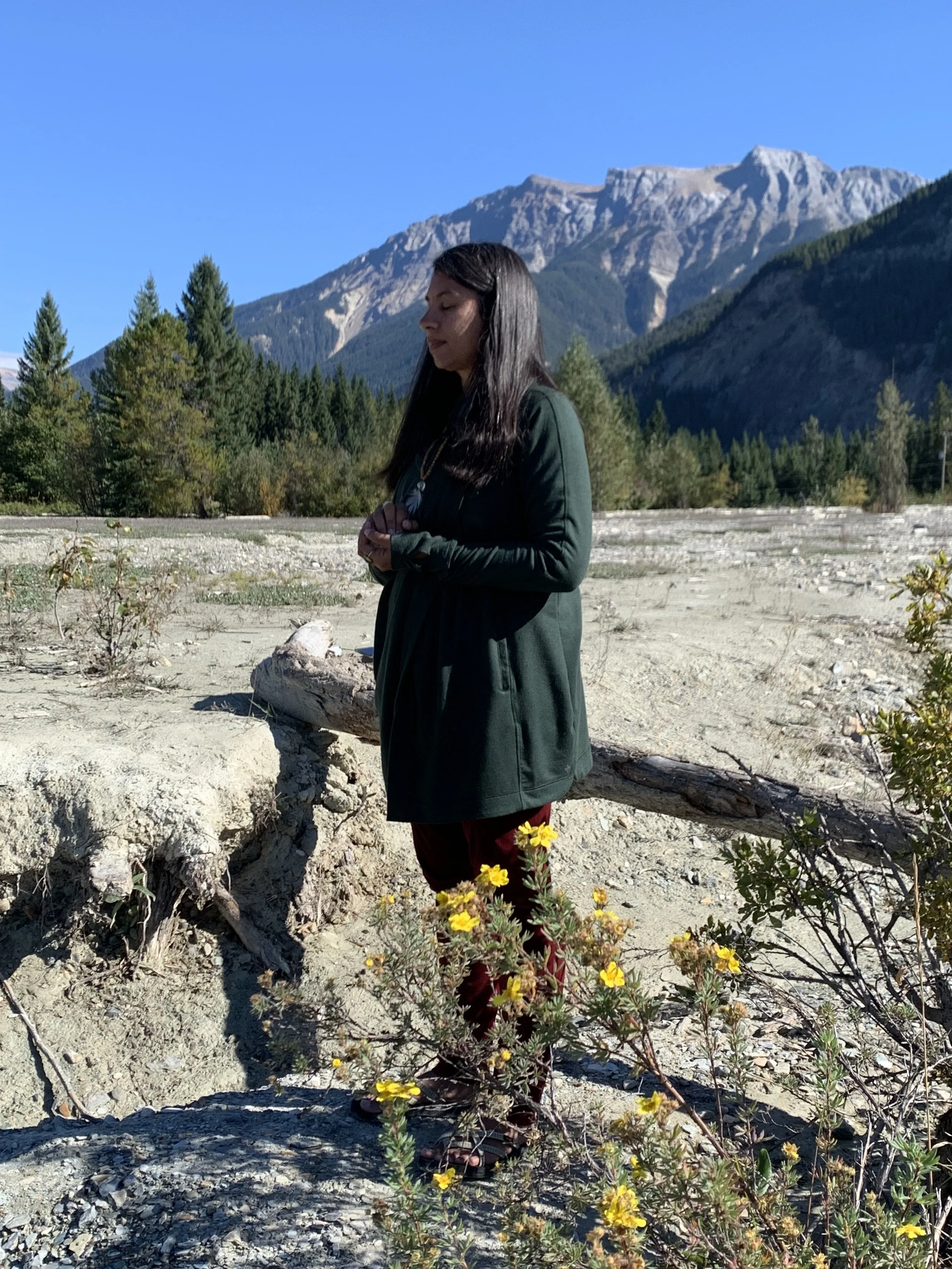 Reverend Clarissa standing in the mountains doing a sacred ritual and ceremony with trees and yellow flowers around her