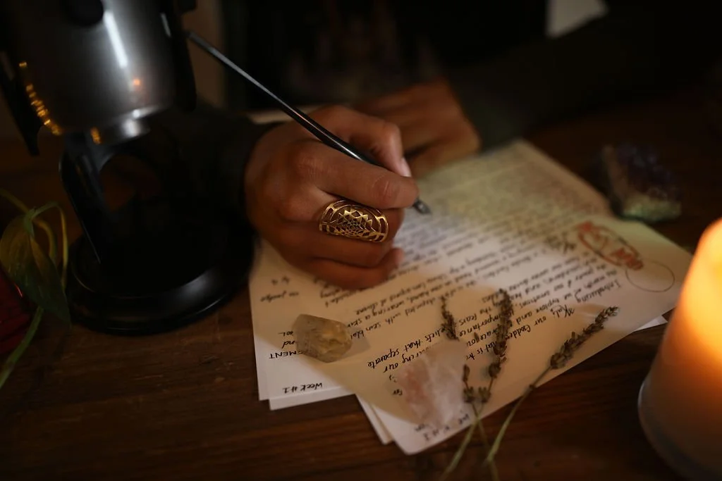 Clarissa's hand with a Sri yantra ring, writing channeled guidance for spiritual growth, with crystals and herbs on the pages