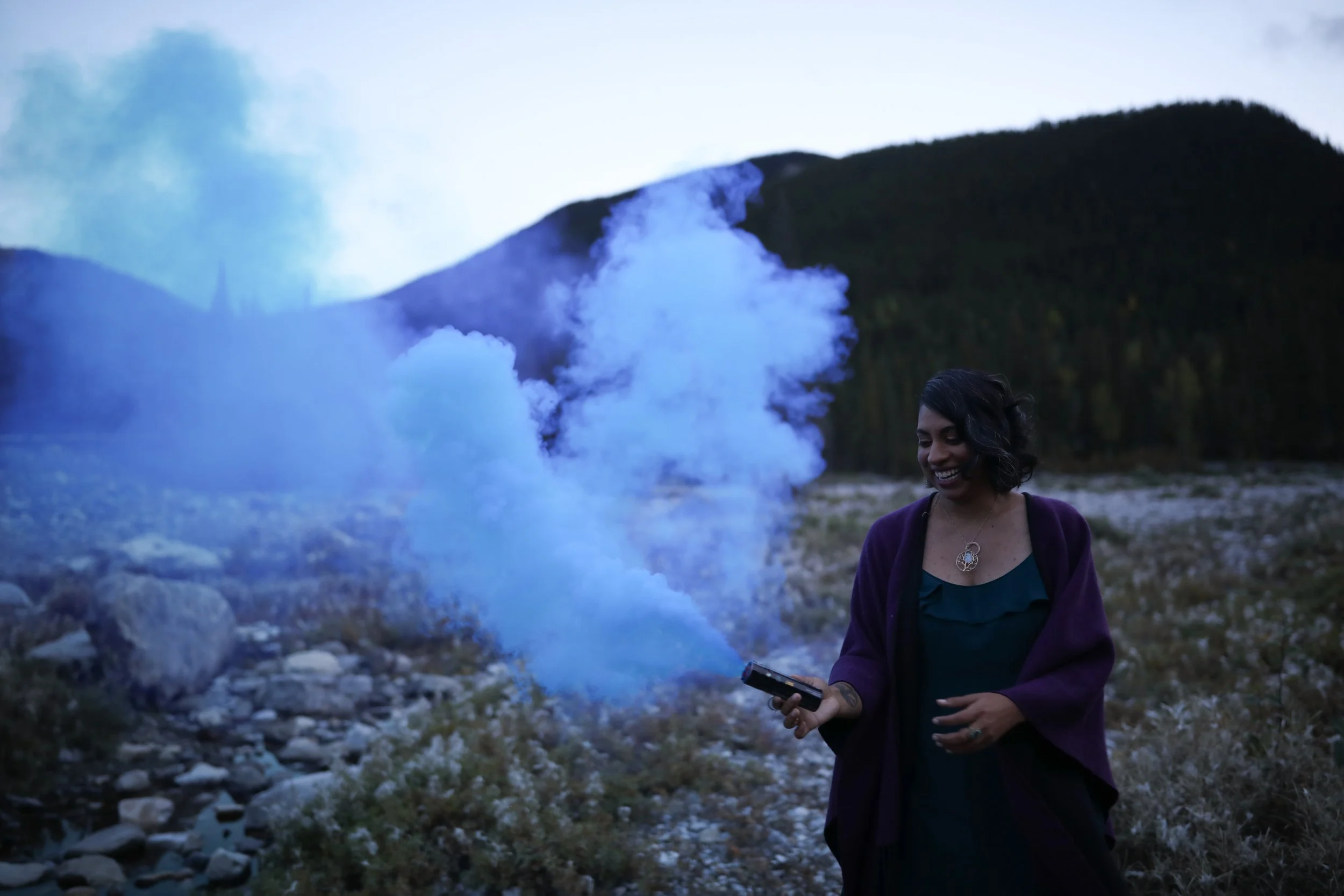 Clarissa laughing in the wildflowers with a mountain behind her, holding a sacred smudge that has blue smoke billowing in a heart shape.