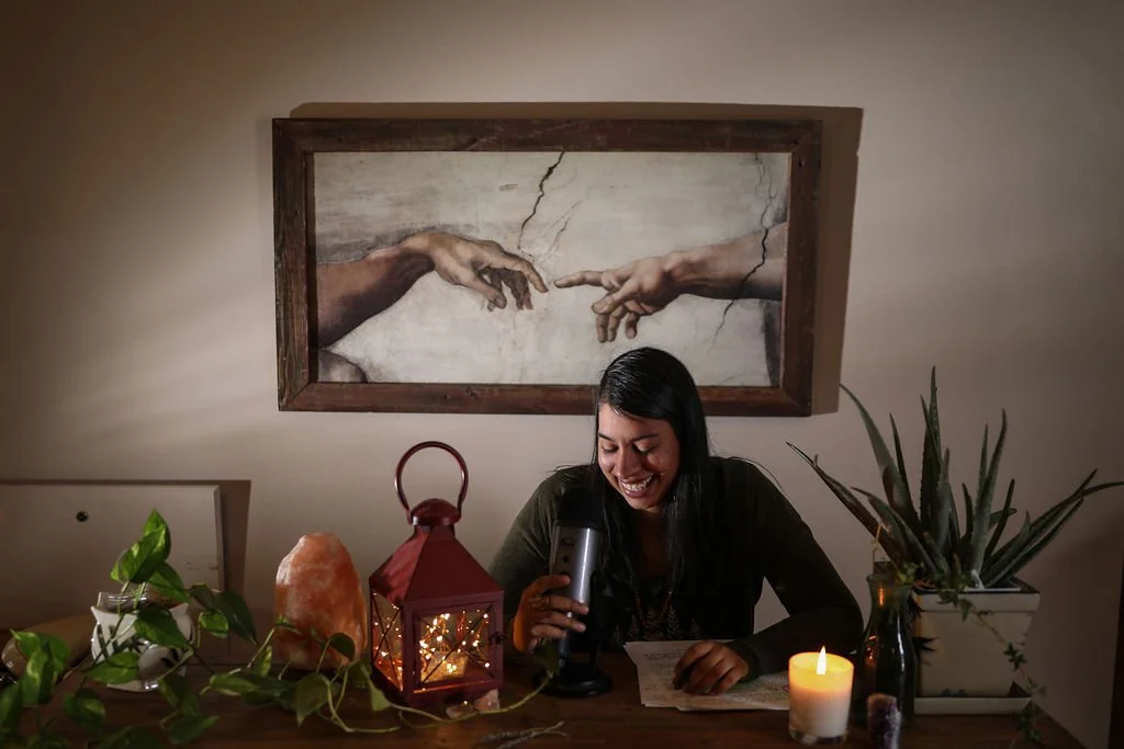 Reverend Clarissa sitting at a desk speaking into a microphone recording readings, there is a lantern, lit candle and salt lamp on the desk
