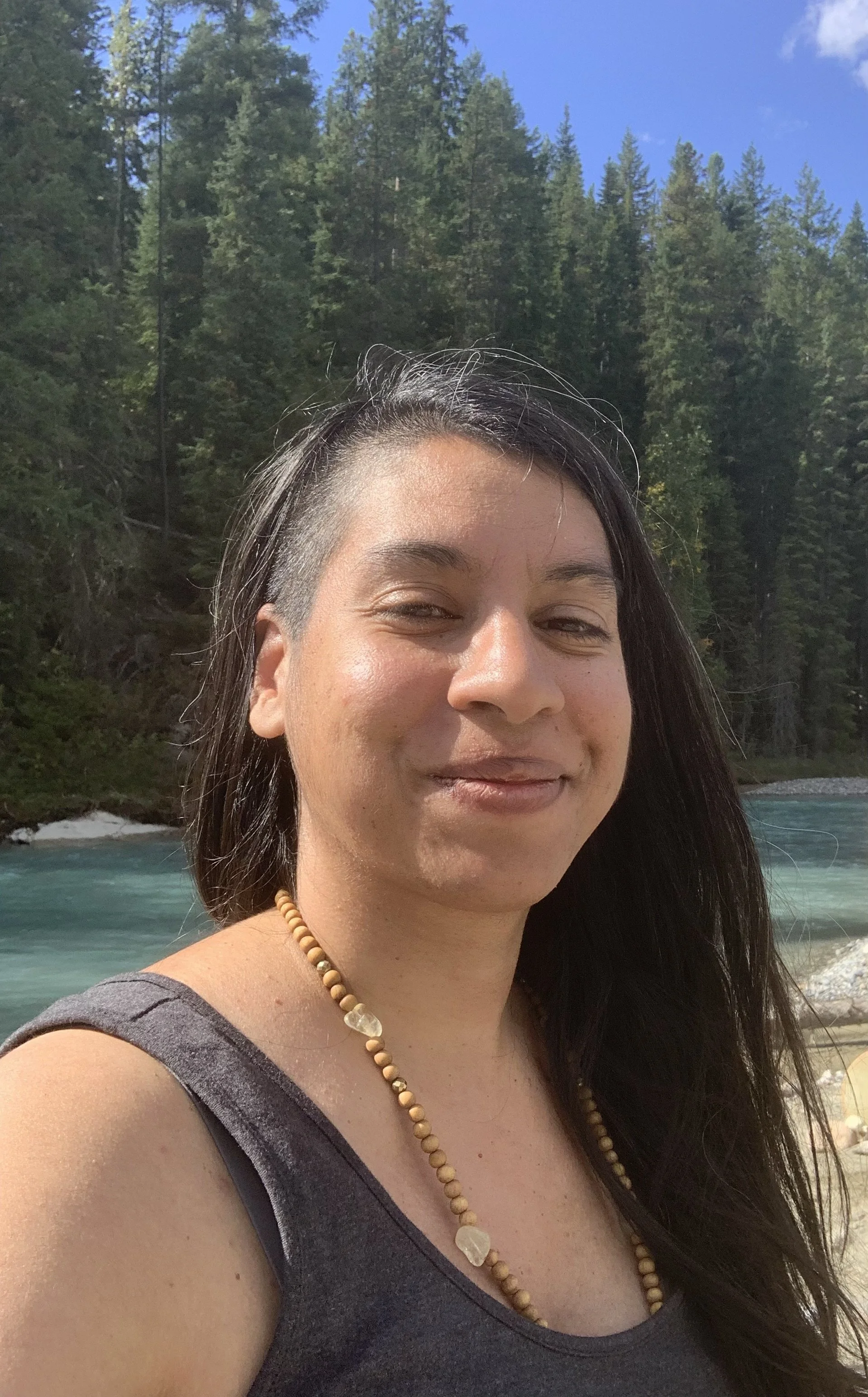 Reverend Clarissa standing in front of a river near a forest of pine trees. she is wearing a citrine mala necklace and a smirk